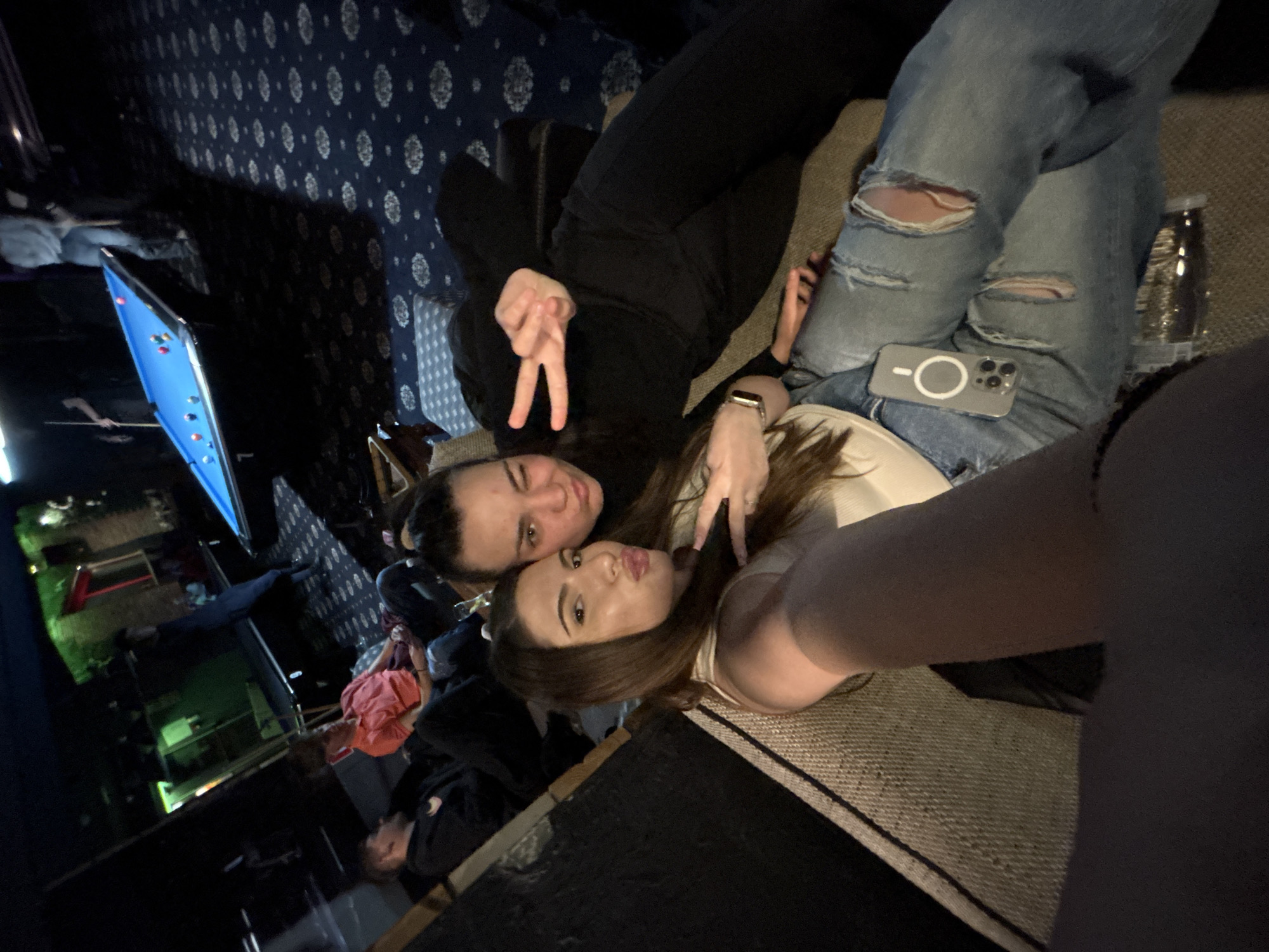 Two young women pose for a selfie indoors. One is wearing a black hoodie and makes a peace sign. The other has long brown hair and wears a white top, also making a peace sign. A smartphone rests on a pair of ripped jeans. A bottle sits beside the jeans. A billiards table and other people are visible in the background. The location is unspecified and there is no information to indicate the image was taken in Plovdiv, Bulgaria.