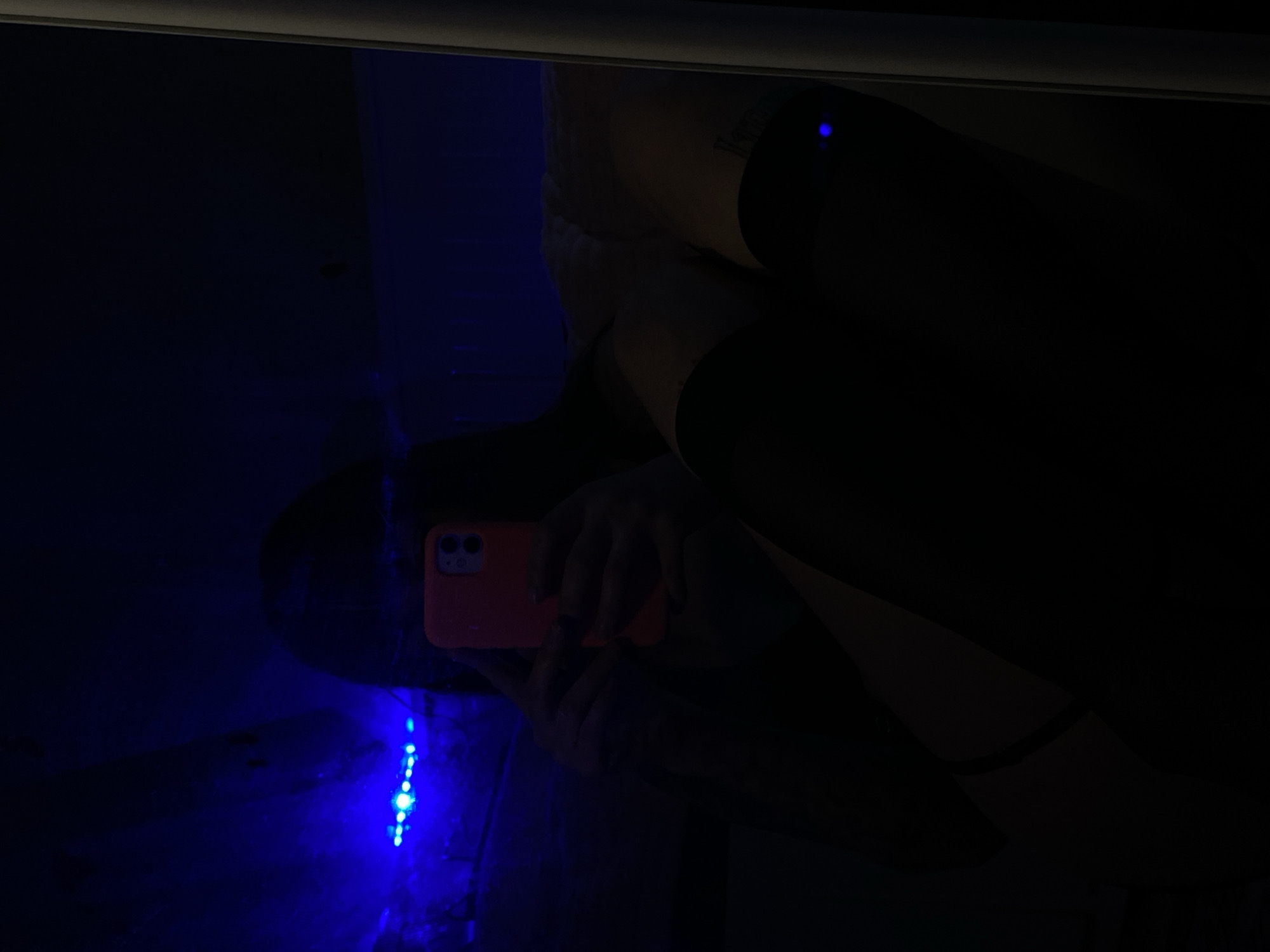 A reflection in a dark room reveals a person holding a phone with a coral-colored case. The phone is positioned to take a picture, possibly a selfie. The person is wearing dark stockings and what appears to be a lace garment. In the reflection, a string of blue LED lights is visible. The location, Votkinsk, Russia, is not determinable from the image.