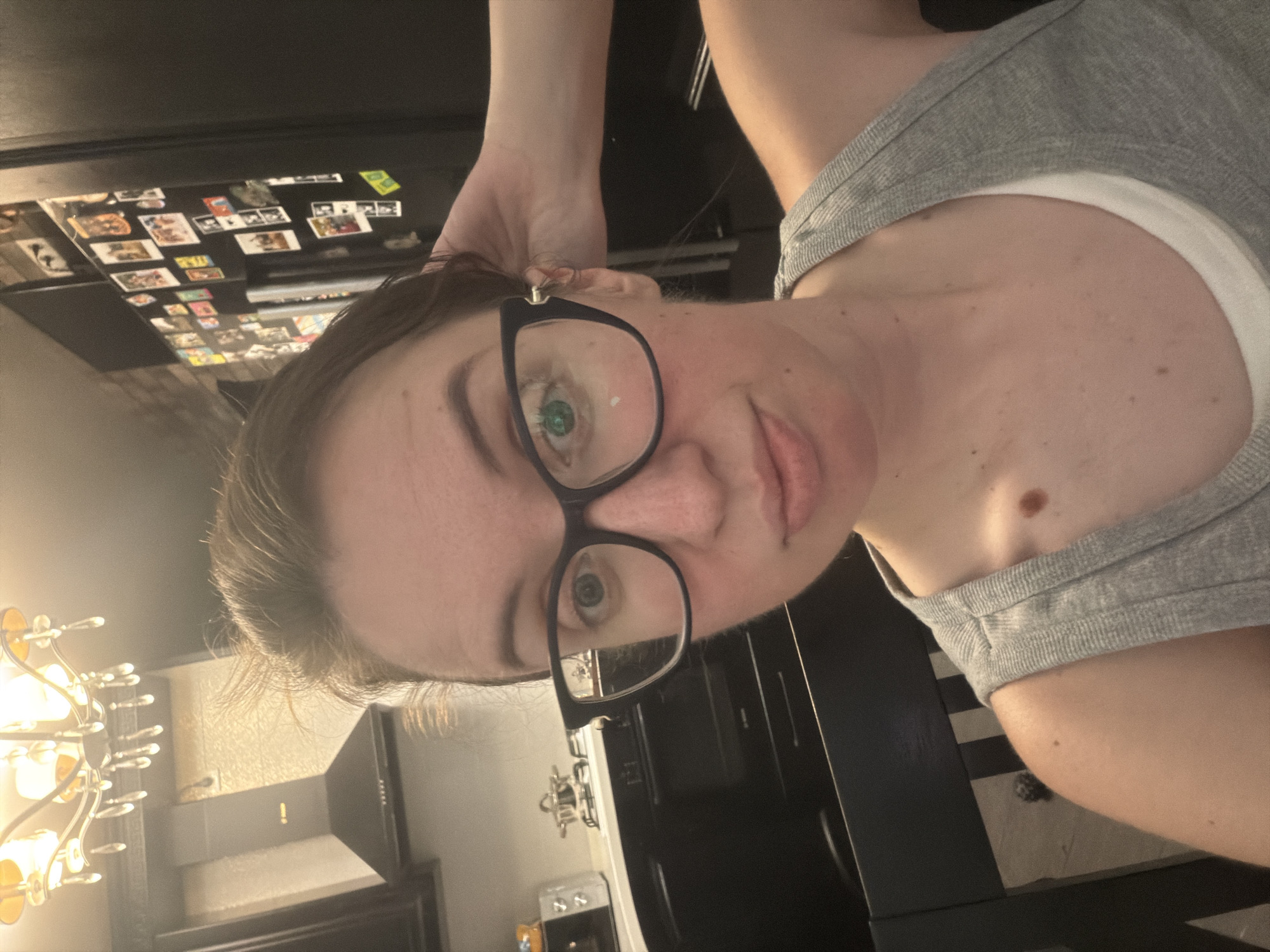 A medium shot captures a woman with brown hair and glasses posing in a kitchen. She is wearing a grey tank top over a white undershirt. The woman's left hand is raised to her head, touching her hair. In the background, a kitchen scene unfolds, featuring a stovetop with a pot, a range hood, a refrigerator adorned with photographs, and a chandelier overhead. The image appears to have been taken indoors, possibly in a residential kitchen, as indicated by the domestic setting and personal decorations. There is no information to suggest the location is the city of Bila Tserkva, Ukraine.
