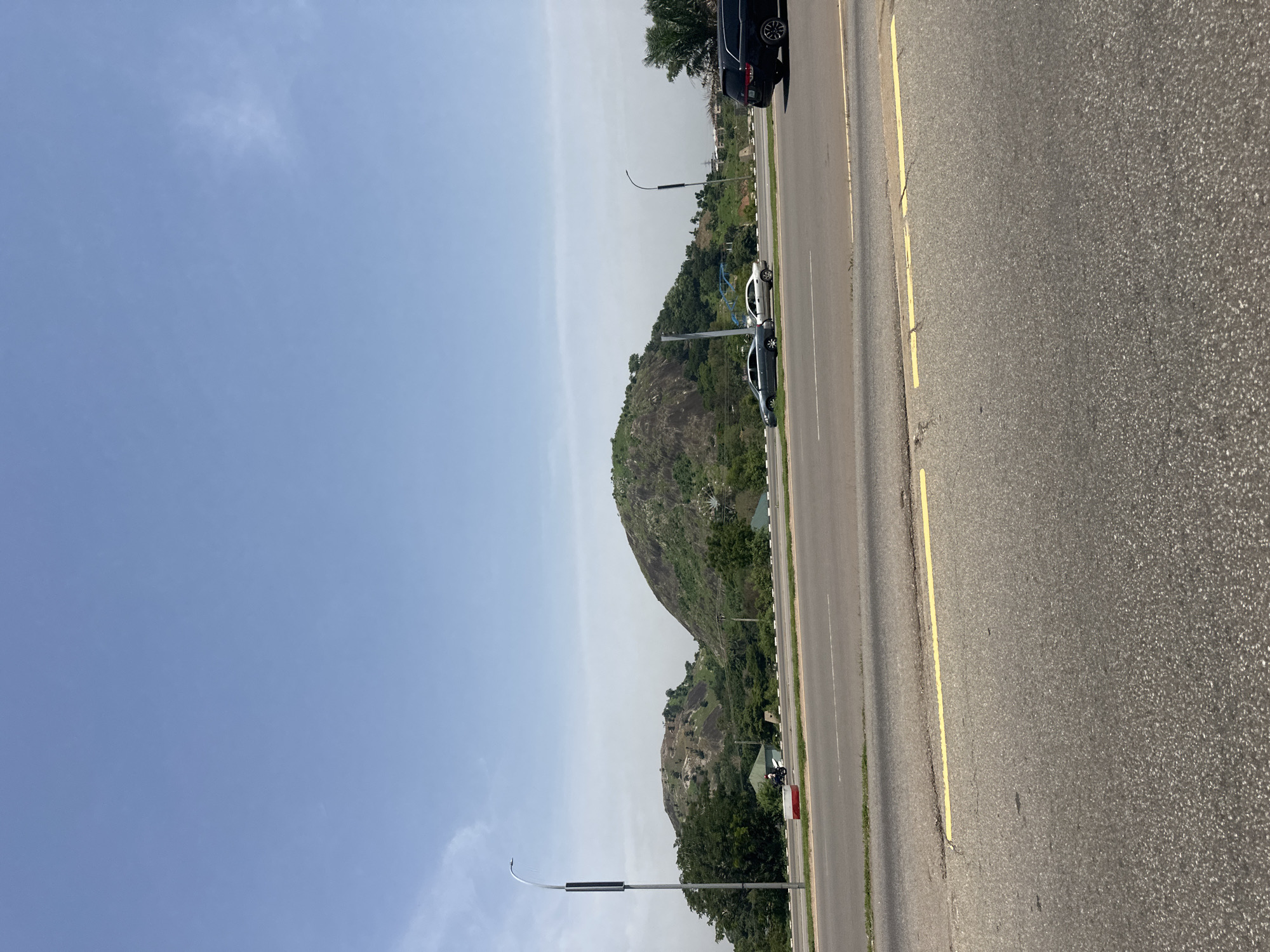 This is a landscape photograph taken in Abuja, Nigeria. The scene features a road in the foreground with a yellow line marking part of the lane. Beyond the road, a grassy area transitions into rolling hills covered in rocks and vegetation. Two cars, a silver sedan and a white sedan, are parked on the side of the road in the middle ground. A dark-colored SUV is visible further down the road on the right. In the background, the landscape is dominated by large, rounded hills with rocky surfaces and patches of green. A tall, slender street lamp with a dark rectangular panel is positioned on the left side of the road. The sky is a clear, pale blue with a few wispy clouds. The overall impression is of a bright, sunny day in an area with natural geological formations.