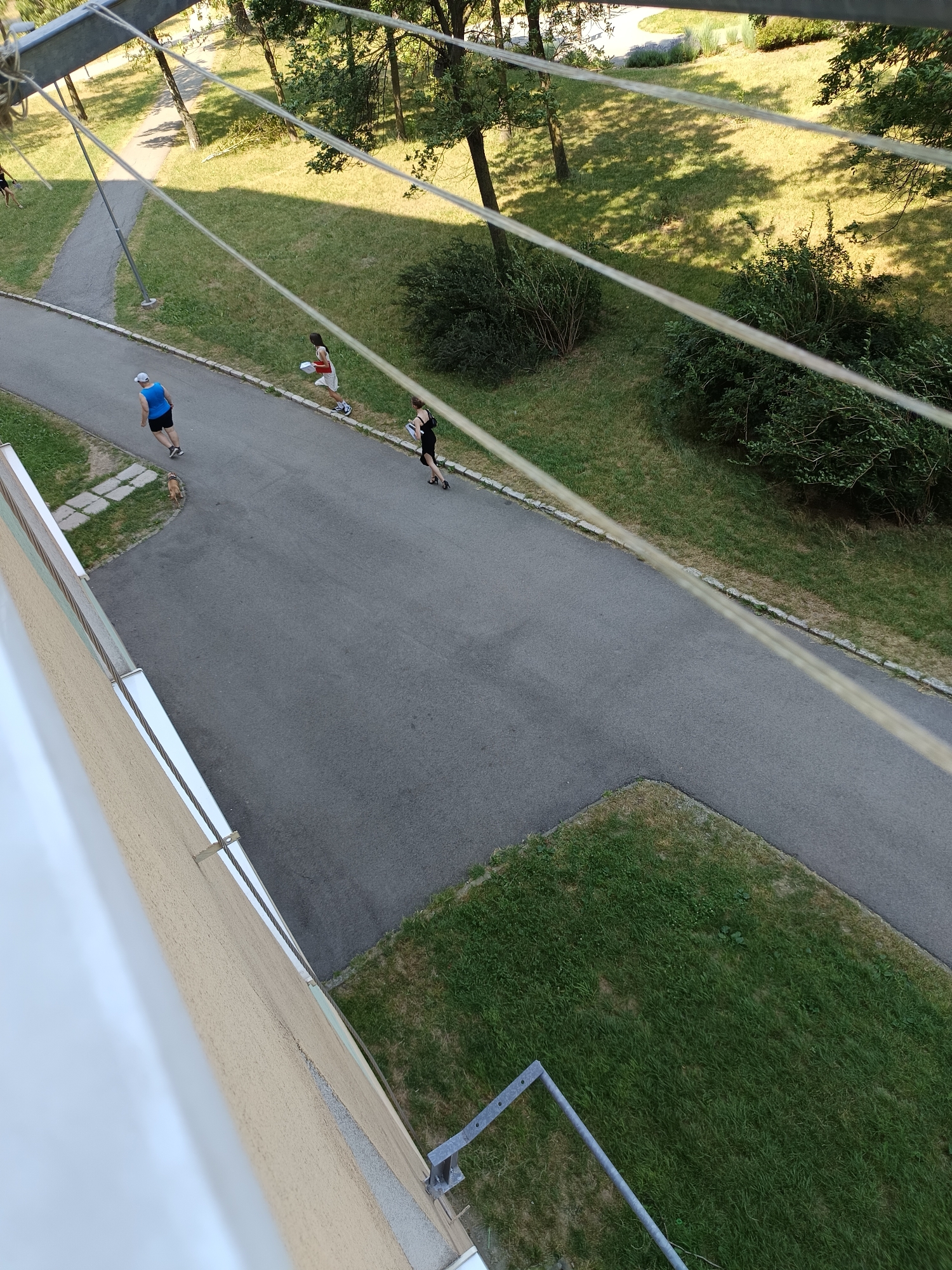 The image shows an aerial view of a paved walkway or road in a park-like setting, likely in Košice, Slovakia, with a building in the foreground. Three individuals are visible on the walkway. The person furthest left is wearing a blue tank top and black shorts, jogging with a small brown dog beside them. To their right, a person in a white top and shorts is walking with a white object. Further ahead, a person in a black outfit is jogging. The walkway is bordered by grass and trees, with shadows cast on the ground from the trees. The building in the foreground has a textured, light-colored facade and appears to be a residential building with balconies or windows. The overall scene suggests a daytime activity in an urban green space.