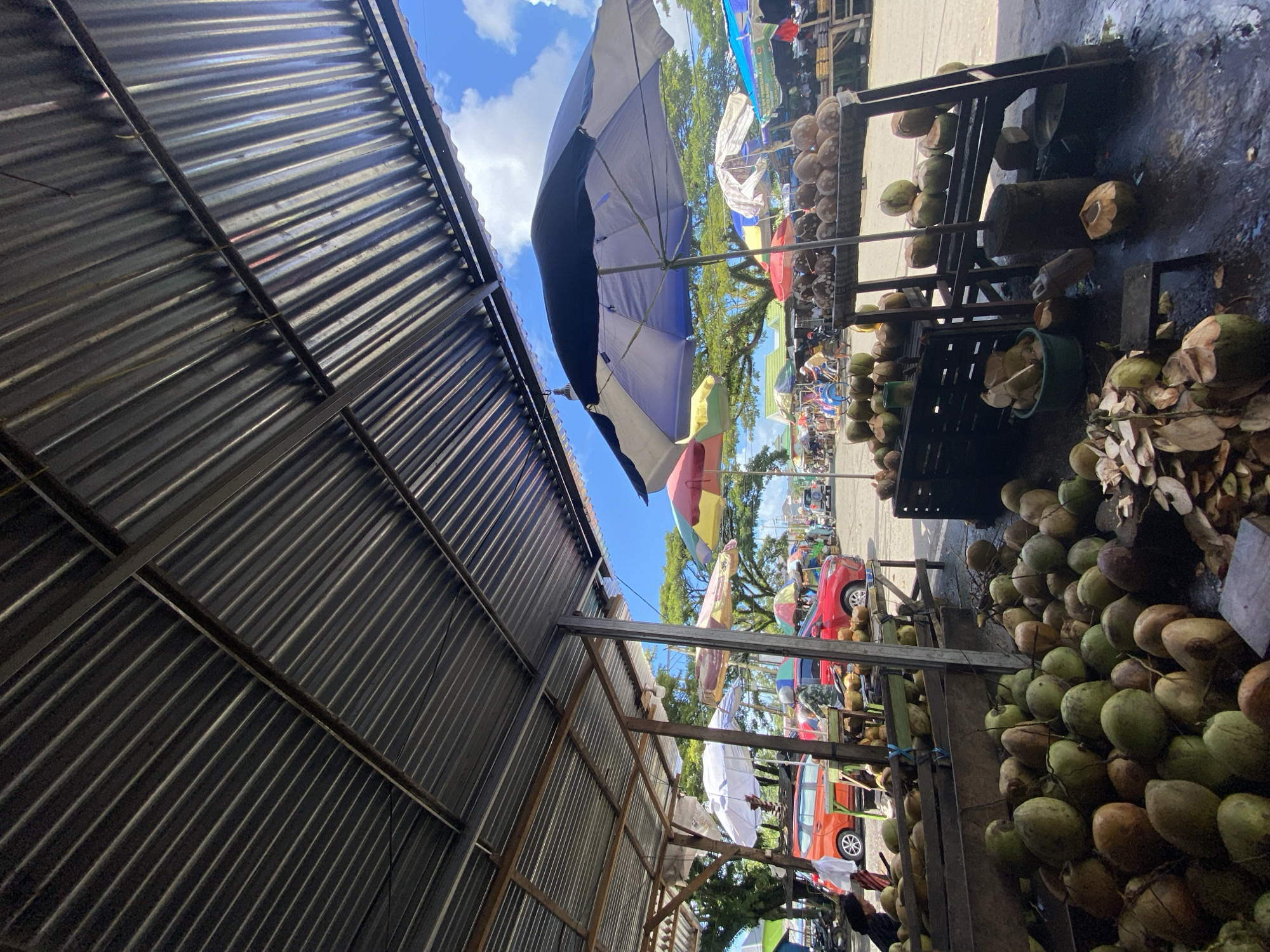 This is an outdoor market scene in the city of Ta', Indonesia, under a corrugated metal roof. In the foreground, a large pile of coconuts is displayed in a wooden bin and on the ground. Some of the coconuts are whole, while others are cut open, revealing their white flesh and liquid. To the right, a tiered wooden shelf holds more coconuts, some stacked and some halved. Several umbrellas, in various colors including blue, white, red, yellow, and green, are set up throughout the market, providing shade. Behind the coconut stalls, several vehicles, including an orange car and a red car, are parked. In the background, more market stalls and trees are visible, and people can be seen moving about. The sky is blue with scattered white clouds. The ground appears wet and reflective in some areas.
