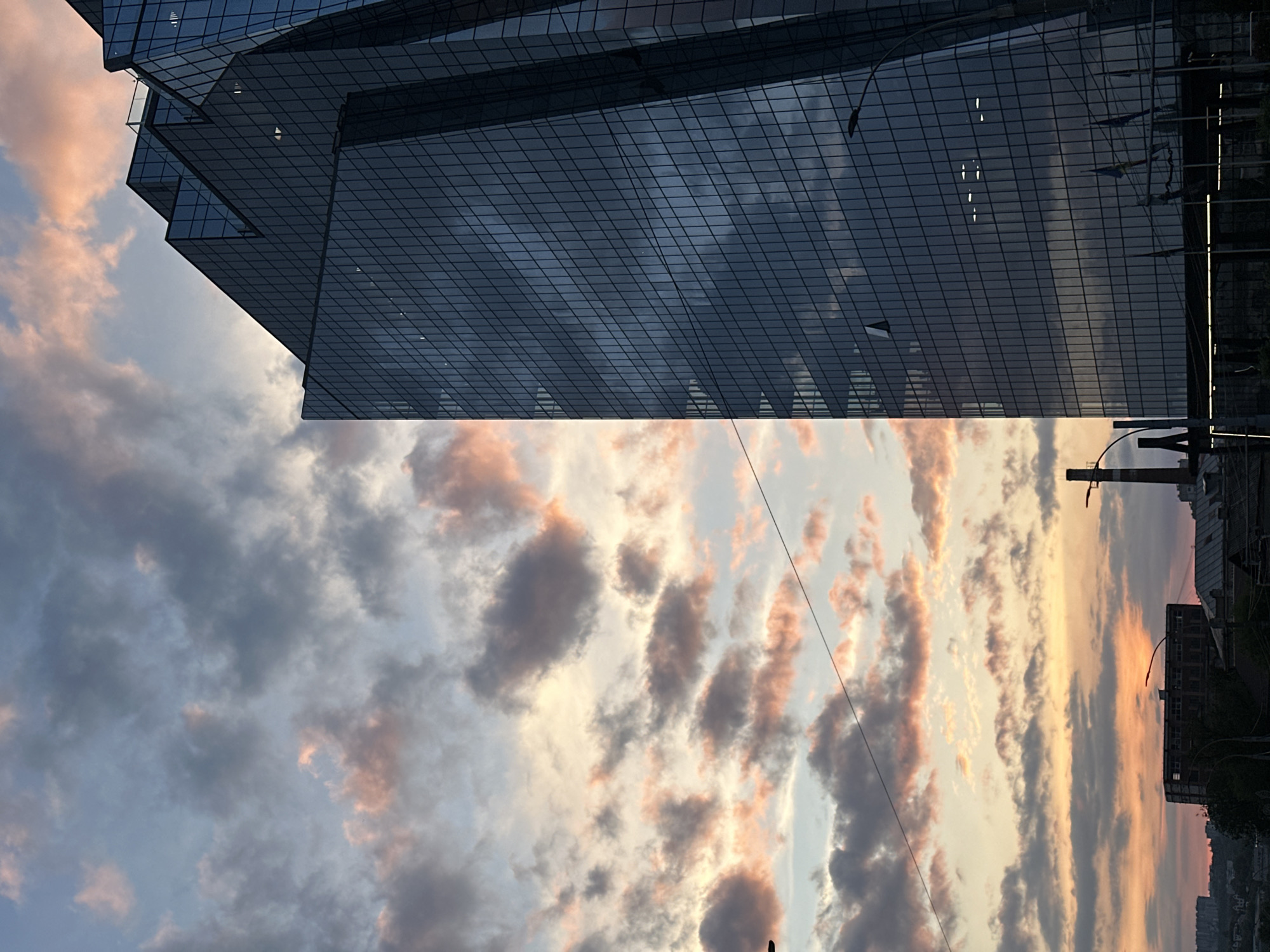 The image shows a low-angle view of a building against a sky with clouds at sunset. The building appears modern, with a glass facade consisting of a grid of rectangular windows. The sky is partially filled with clouds that are tinged with pink and orange hues, indicating a sunset.
The location is likely in Chișinău, Moldova, because the building is the Radisson Blu Leogrand Hotel, located in the city center of Chișinău. The street lamps and power lines are visible. The other buildings are visible, and the overall environment suggests an urban setting. No people are visible in the image.