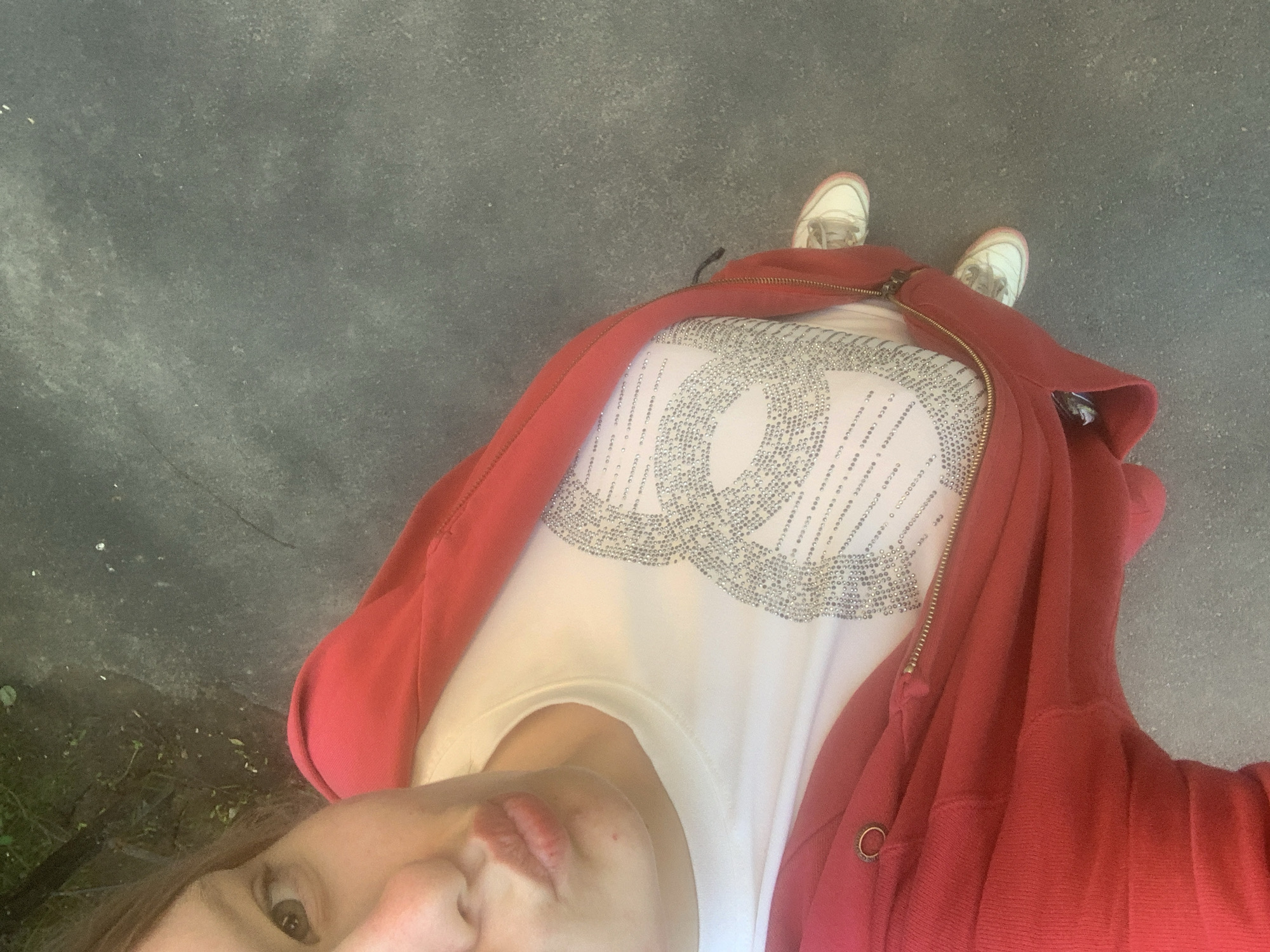 An overhead, high-angle selfie depicts an individual outdoors in Ladozhskoe Ozero, Russia. The person, with fair skin, is partially visible, showing one brown eye looking directly at the camera and pursed lips. A small red blemish is present on the chin. Brown hair is visible on the upper left. The individual wears a white t-shirt featuring a large, intricate design composed of numerous small, reflective studs or rhinestones, forming a stylized, interlocking pattern that includes elements resembling dots or micro-text. A red zip-up hoodie or jacket, partially unzipped with a brass-toned zipper, is worn over the t-shirt. A brass ring detail is visible on the jacket's left side. White sneakers with light brown laces and light pink trim on the soles are visible at the bottom. The ground is a textured grey surface, likely concrete or asphalt. The upper left background shows out-of-focus green foliage and dark ground, suggesting a natural element adjacent to the paved area.