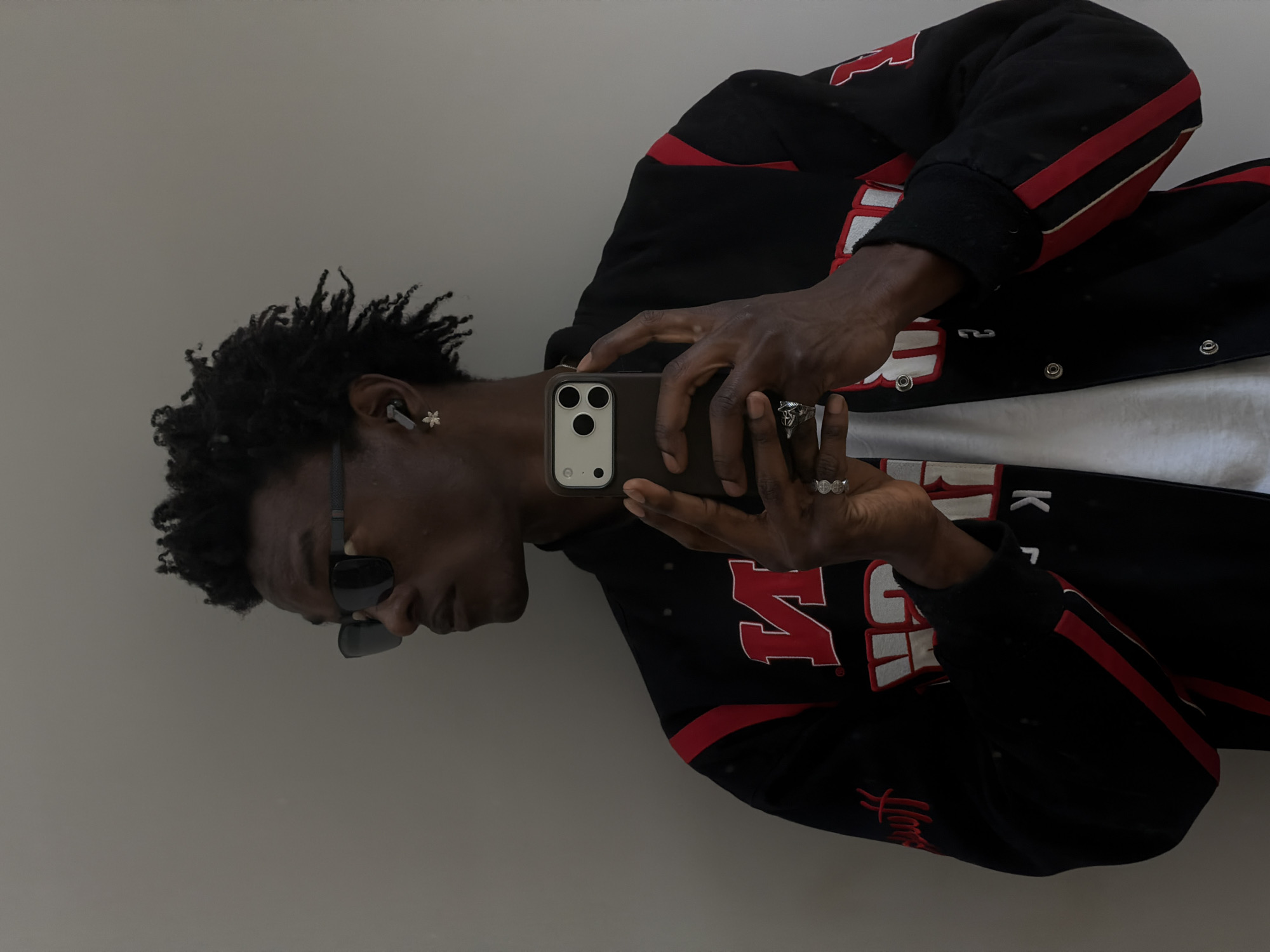 A man is holding a phone, presumably taking a selfie. He's wearing sunglasses and a black jacket with red and white accents, along with a white shirt. He is also wearing an earpiece. The background is a plain, light gray surface. There is no indication of the location being in London.