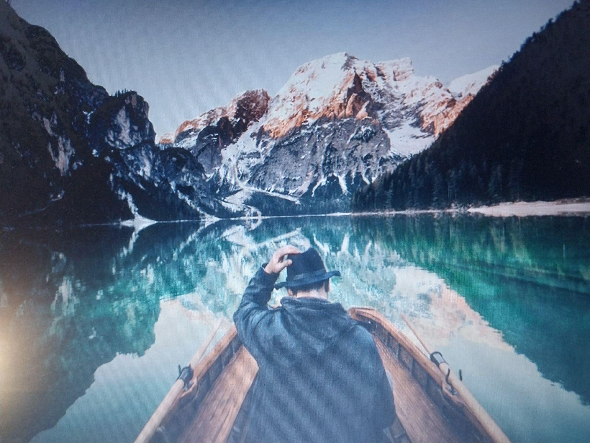 A man in a black fedora and a dark jacket is sitting in a wooden boat. He is facing away from the camera, looking towards snow-capped mountains and a lake. The mountains are covered in snow and have rocky outcrops. The lake is calm and reflects the mountains and the sky. The sky is clear and a pale blue. The boat is in the foreground, with its wooden hull visible. The scene appears to be a landscape, likely in a mountainous region. The man is touching his hat, perhaps to adjust it or to acknowledge the view.