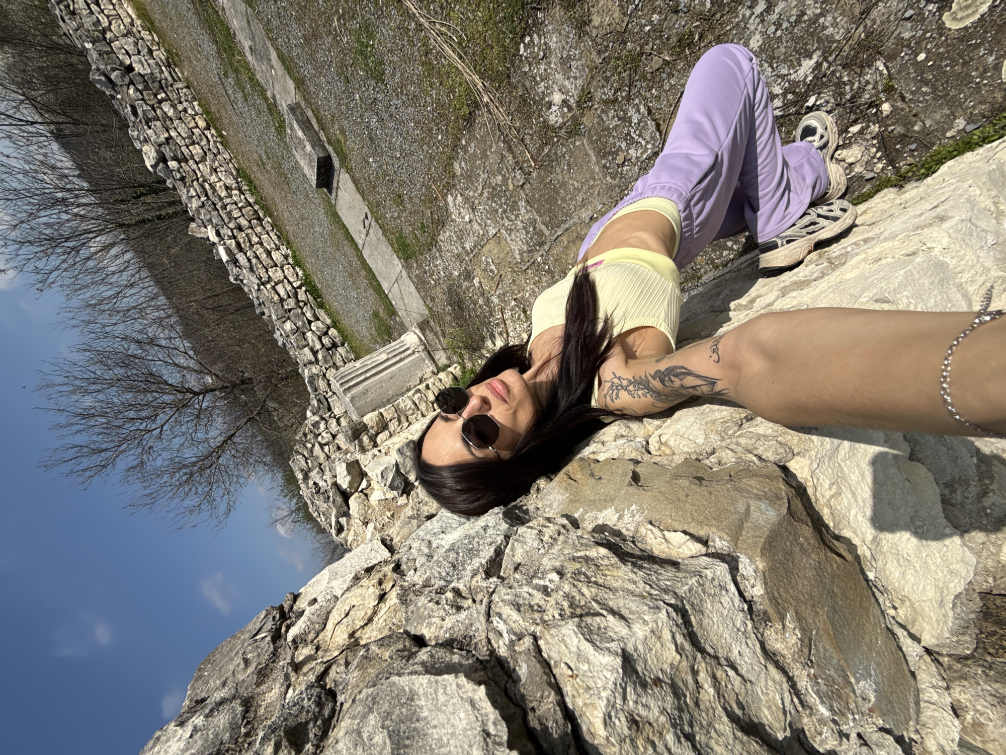 A woman lies on a stone structure in Pleven, Bulgaria. She wears sunglasses, a yellow top, and purple pants. She has dark hair and a tattoo on her arm. The background includes a stone wall, sparse trees, a gravel path, and a blue sky.