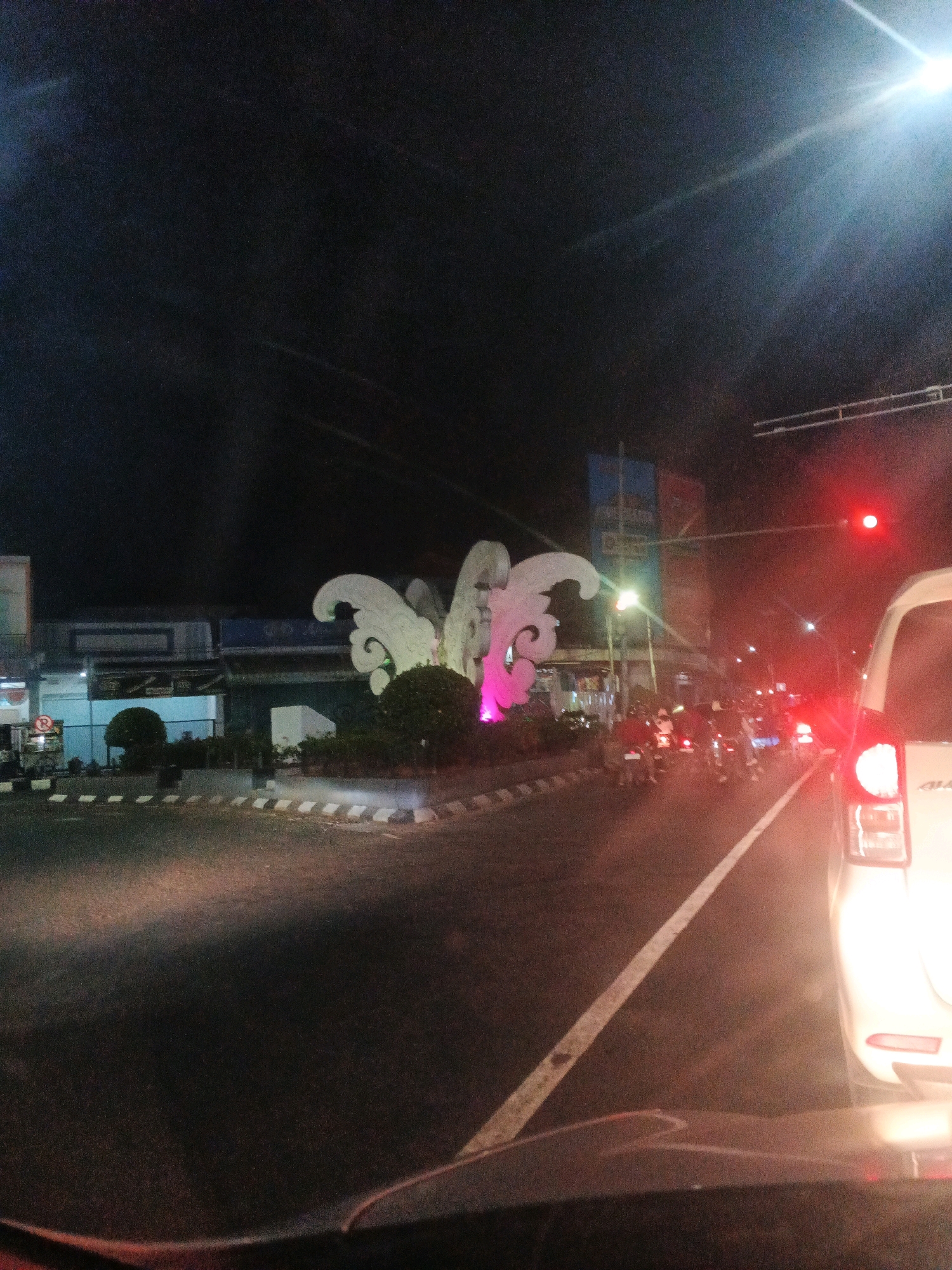 At night in Jatisari, Indonesia, a street scene unfolds. A white vehicle, viewed from the rear, occupies the right side of the frame. Beyond it, a traffic light displays a red signal, illuminated by a bright red glow. A line of motorcycles waits at the intersection, their headlights cutting through the darkness.
In the center of the image, a large, decorative structure resembling a butterfly or a stylized flower, illuminated with pink and white lights, stands amidst lush green bushes. This landmark is situated on a traffic island bordered by a white and black checkered pattern along the road.
To the left, a building with storefronts is visible, featuring signage and lights. The street is wet, reflecting the surrounding lights, suggesting recent rain or general humidity. The sky is dark, with a bright light source, possibly a streetlamp or a strong distant light, creating a lens flare effect in the upper right quadrant. The overall atmosphere is that of a typical urban night scene, with moderate traffic and illuminated public spaces.