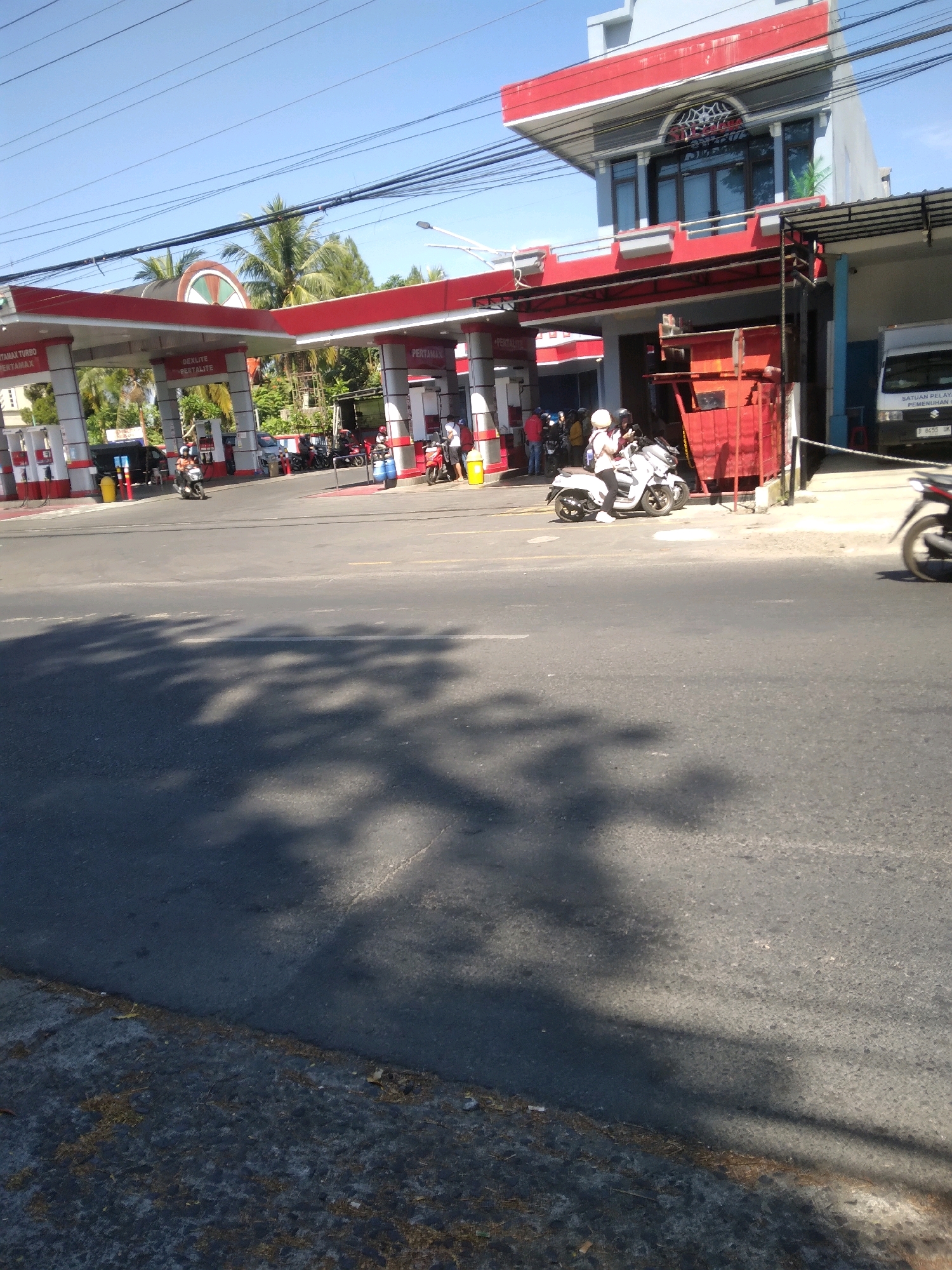 The image depicts a street scene outside a Pertamina gas station in Grendeng, Indonesia. The gas station features red and white striped pillars supporting a red roof. Several motorcycles are present, including one parked in front of the station and another being ridden by two individuals. People are seen interacting near the gas pumps and on the sidewalks. The environment appears to be sunny with clear blue skies and shadows cast by trees on the asphalt road. A white truck with Indonesian license plates is parked near the station.