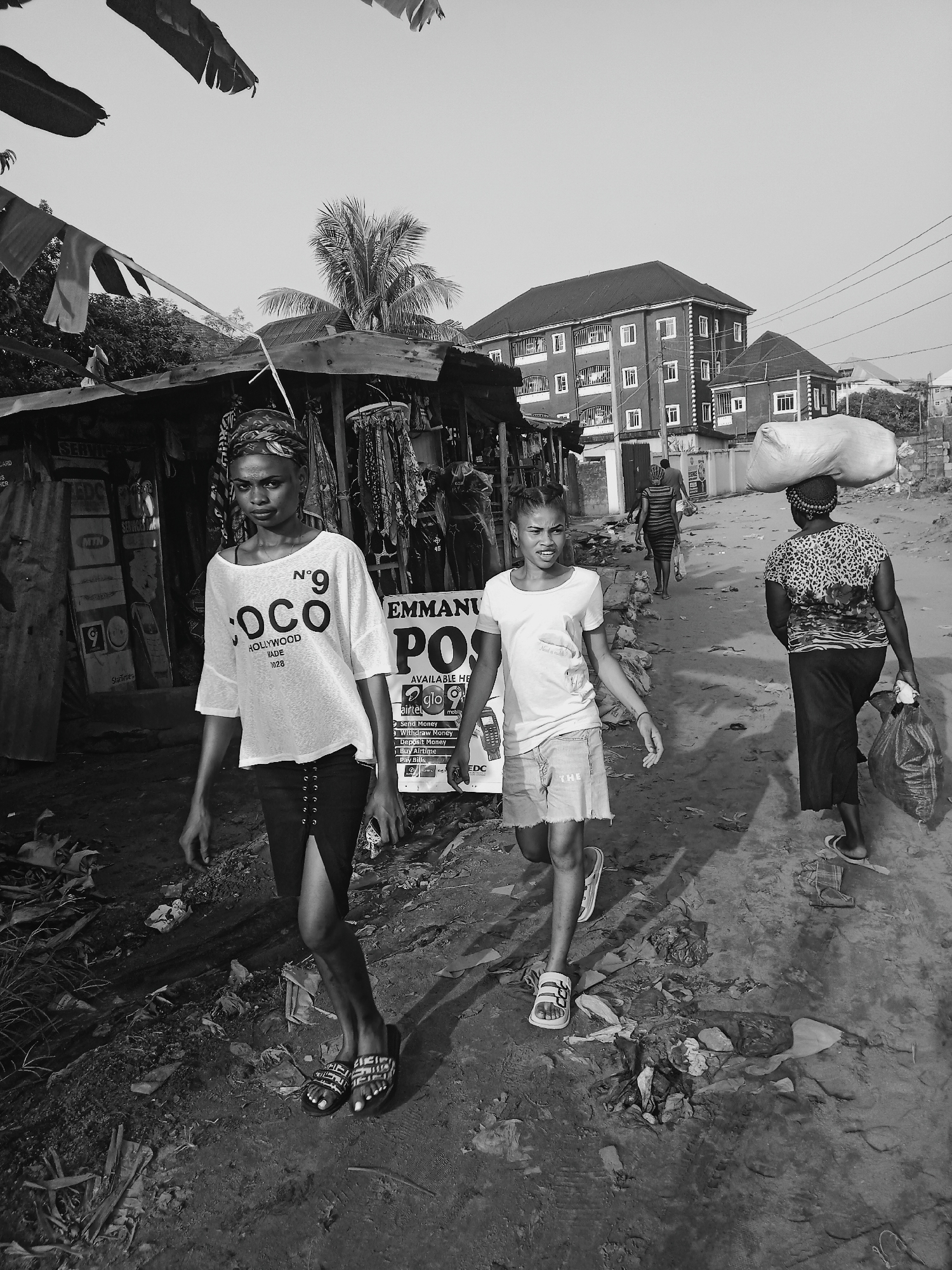 Captured in Onitsha, Nigeria, a grayscale photograph presents a street scene marked by a rough, littered ground. A young woman with a head wrap, wearing a "Coco" t-shirt and skirt with sandals walks at the forefront. A younger girl, clad in a plain t-shirt, denim shorts, and white sandals, accompanies her. In the background, a woman balances a large white sack on her head while carrying a bag. A partially visible commercial stall flanks the left side, with advertisements for mobile money services. Buildings, including a three-story residential structure, line the backdrop. Overhead, electrical wires crisscross the sky. The scene conveys a sense of everyday life amidst the challenges of urban poverty.