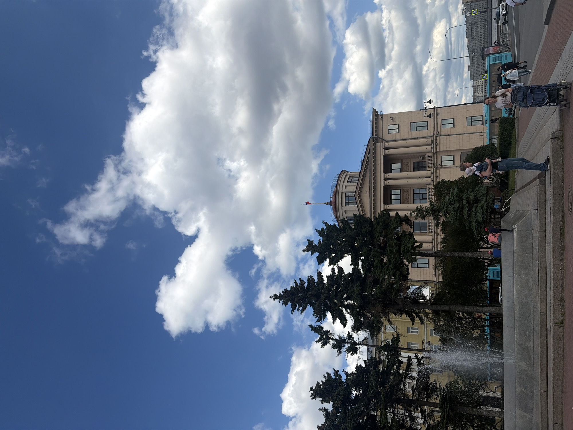 This is an eye-level, medium shot of a waterfront area in Saint Petersburg, Russia, on a cloudy day with bright blue sky. The dominant structure is a light brown, neoclassical building with a circular dome, adorned with columns and a flag at its peak. In the foreground, a large stone fountain sprays water upwards. Several tall, dark green evergreen trees are positioned to the left of the fountain and partially obscure some of the buildings behind them.
To the right of the fountain, a man is holding a baby, dressed in denim overalls. A woman is pushing a stroller with a child in it. Further in the background, a bright blue bus is visible, and beyond that, the Neva River can be seen, with a distant city skyline on the other side. Pedestrians are walking along the waterfront promenade, and cars are visible on the street. The overall scene depicts a public space within the city, likely a park or plaza, enjoying a partially sunny day.