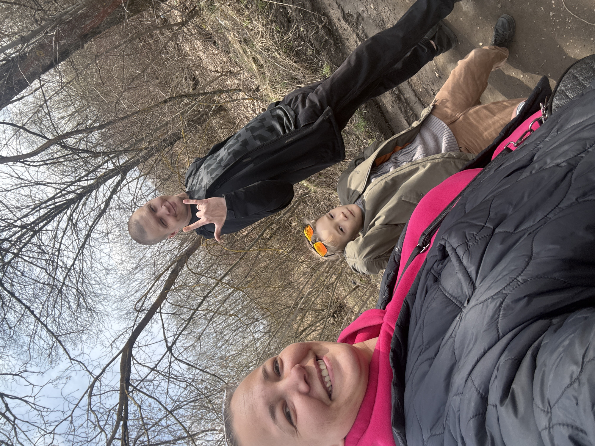 Three people are in a wooded area on a sunny day in Dmitrov, Russia. The photo, a selfie, shows a woman smiling with trees in the background. Behind her, a man makes a "rock on" hand gesture. A child with orange sunglasses is lying down as well. Bare trees surround the group. The ground is dirt.