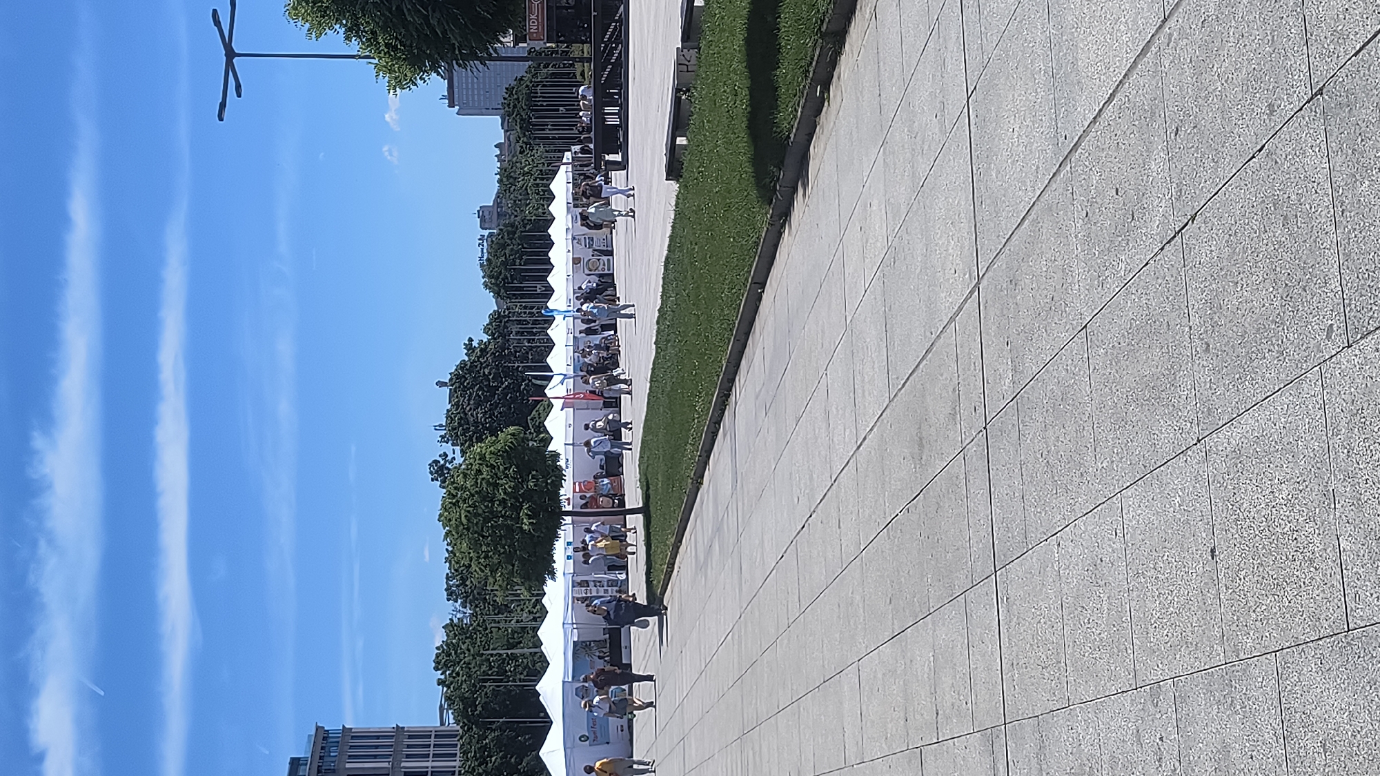 The image shows a long row of white vendor tents set up along a paved pedestrian walkway in Sofia, Bulgaria, likely at a public event or market. People are seen walking along the walkway, interacting with the vendors. The walkway is bordered by a strip of green grass and trees. In the background are buildings and a clear, blue sky with wispy clouds. A tall light post stands in the upper left corner of the image. The location appears to be near the National Palace of Culture (NDK).