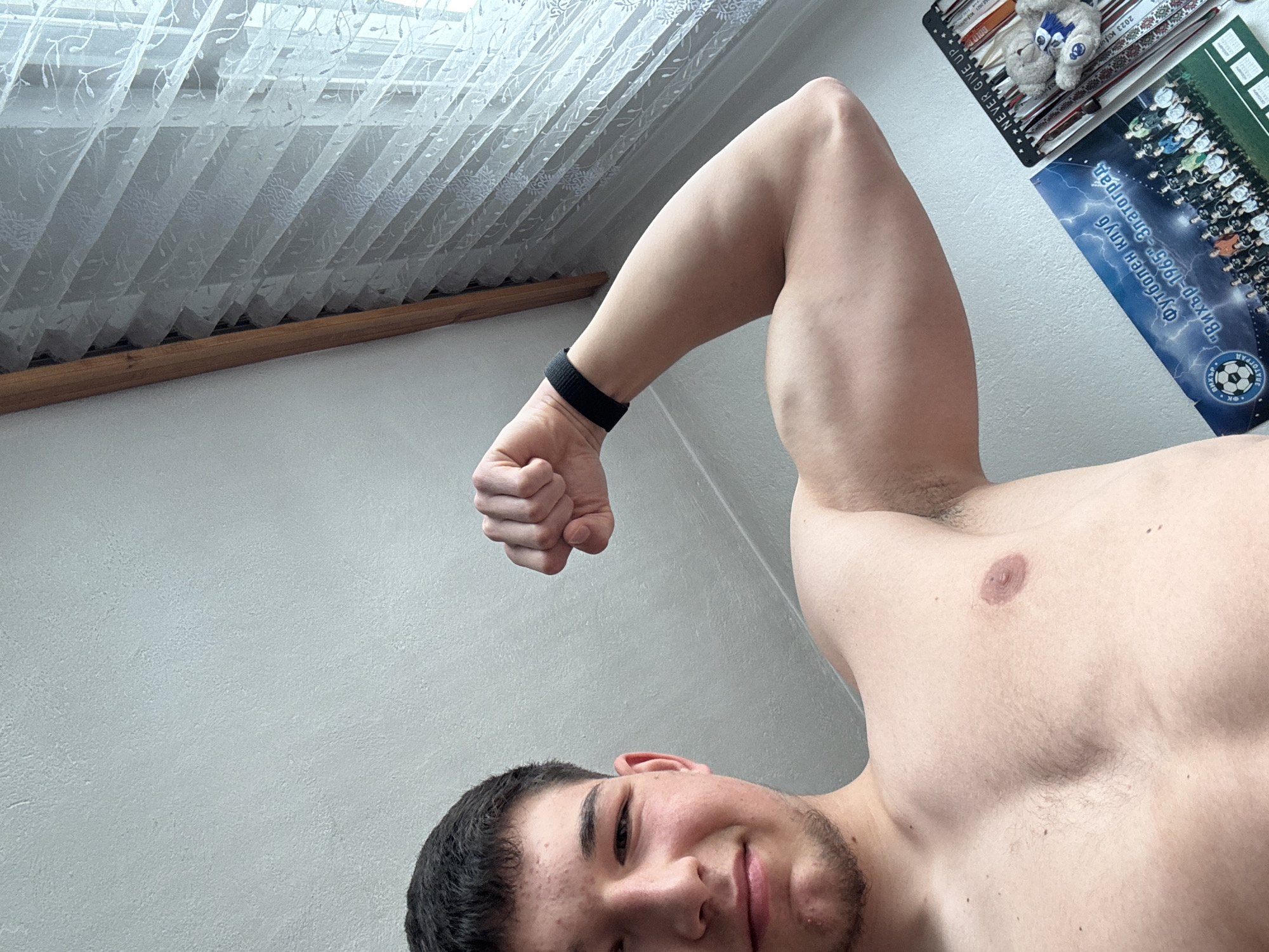 A shirtless young man with dark hair poses for a selfie, flexing his right bicep. He wears a black wristband. The background is a light grey wall and a window covered with white sheer curtains. To the right and above him is a desk with a poster featuring a Bulgarian soccer team called "Вихър-1965" (Vihr-1965) from the city of Zlatograd.
