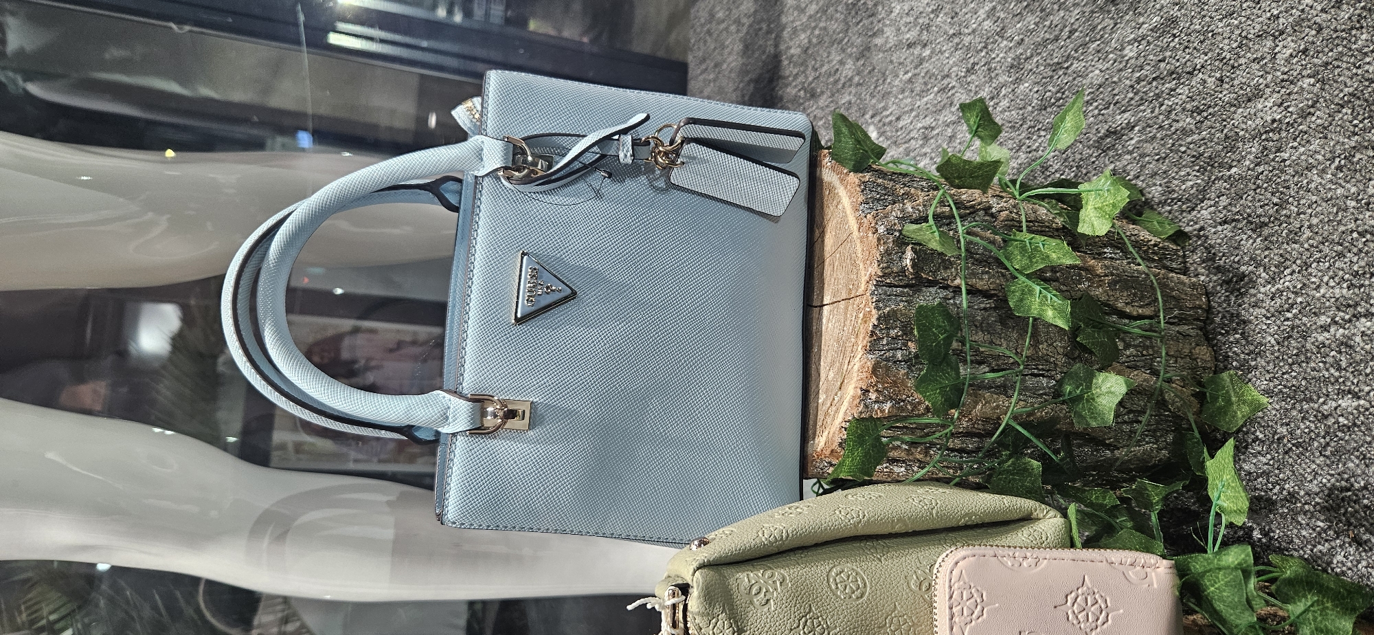 A light blue Guess brand handbag with a textured finish and gold-toned hardware is displayed on a tree stump. The handbag has two top handles and a detachable shoulder strap. Two grey rectangular hang tags are attached to the bag. To the left of the handbag, a portion of a light green handbag with embossed floral patterns is visible. The tree stump is decorated with artificial green ivy leaves. The background is blurred and appears to be a store display with mannequins and glass. The scene is likely located in a retail environment, possibly in the city of Hévíz, Hungary, given the context. There are no people or actions depicted in the image. The lighting suggests an indoor setting, possibly a shop window.