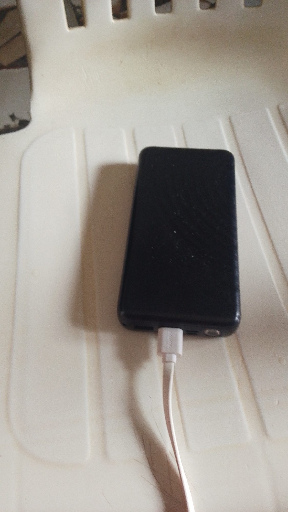 A black, rectangular power bank is plugged into a white charging cable and rests on a textured, light-colored surface. The power bank is positioned vertically and slightly off-center to the right of the frame. The charging cable is connected to the bottom of the power bank and extends downwards and to the right, casting a shadow. The surface appears to be a chair or bench, with parallel raised lines and a slightly uneven texture. The background is a blurry expanse of the same light-colored material, with some darker, indeterminate objects visible at the top left of the image. There are no people or animals in the scene.