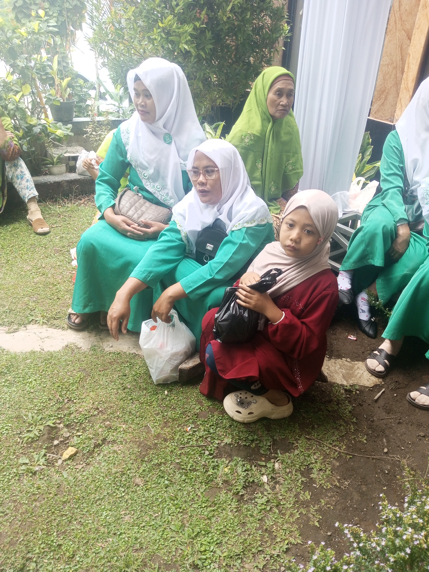 In Klaseman, Indonesia, a group of four females, three adults and one child, are seated outdoors on a patch of grass. The adults are wearing traditional Islamic attire, with two in green dresses and white hijabs, and one in a green hijab. The child is wearing a maroon dress and a light beige hijab, and is holding a black plastic bag. The background consists of greenery and white drapery. The ground is covered with grass and dirt, with a few scattered rocks and a white plastic bag. The scene suggests a casual gathering or event taking place outdoors.