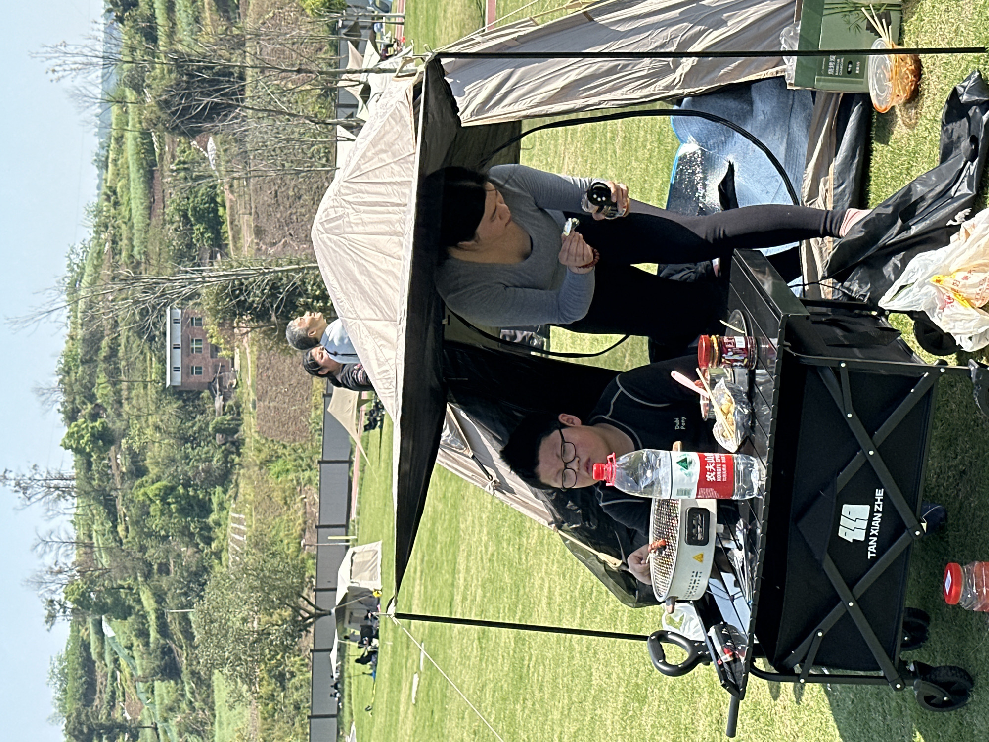 Here is a detailed description of the image:
**General Scene:**
The photo captures a picnic scene, likely in a park or recreational area in Jiangjin, China. There are tents, people, and equipment indicating an outdoor gathering.
**People:**
Two individuals are central in the frame, seemingly seated or resting within the tent structure. Other people are also present.
**Objects:**
Tents: Several tents are set up on the grass. One tent is directly overhead, featuring a canopy.
Picnic Setup: A black wagon with the Chinese characters "TAN XIAN ZHE" on its side appears to be serving as a makeshift table. There are plates with food, water bottles with Chinese labels.
Food: Fried potatoes can be seen on the portable table.
**Actions and Interactions:**
People are preparing food and relaxing. The people look at the camera, posing for the shot.
**Environment:**
Grass: The picnic takes place on a grassy area.
Trees: Trees can be seen in the background.
Buildings: Structures, possibly residential buildings, can be spotted in the background.
Weather: The weather appears to be sunny and clear, based on the visible light and shadows.
Landscape: A rolling landscape with terraces.
Jiangjin China: The Chinese brands indicate that this is located in China. The address and landmarks are not specific to describe further.