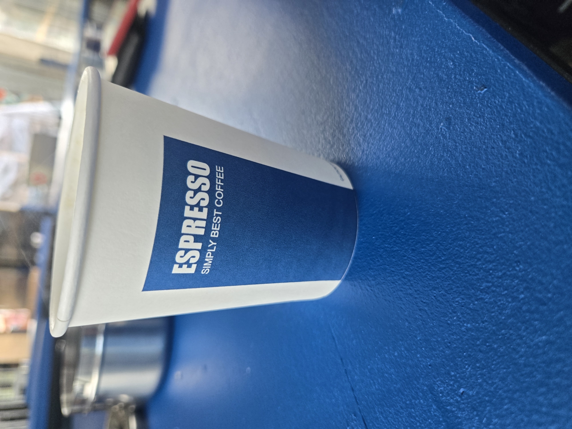The image displays a white paper coffee cup resting horizontally on a textured blue surface. The cup features a prominent blue label with white text. In large, bold capital letters, "ESPRESSO" is printed, with "SIMPLY BEST COFFEE" in smaller capital letters beneath it. The cup's upper rim is visible, appearing clean and white, while the body of the cup is a uniform white.
The blue surface beneath the cup has a distinct, somewhat pebbled texture, suggesting it might be painted wood, plastic, or a similar coated material. There are minor imperfections and variations in the surface's finish. The background is out of focus, showing blurry shapes and metallic reflections. These background elements, which include what looks like a silver, cylindrical part of a machine, hint at an indoor setting such as a cafe, coffee shop, or a commercial kitchen area.
No people are visible in the image, and no specific activity is taking place; the cup is simply depicted resting on the surface. The lighting appears even and indoor, providing no clues about the time of day or external weather conditions. The visible text on the cup is "ESPRESSO" and "SIMPLY BEST COFFEE."