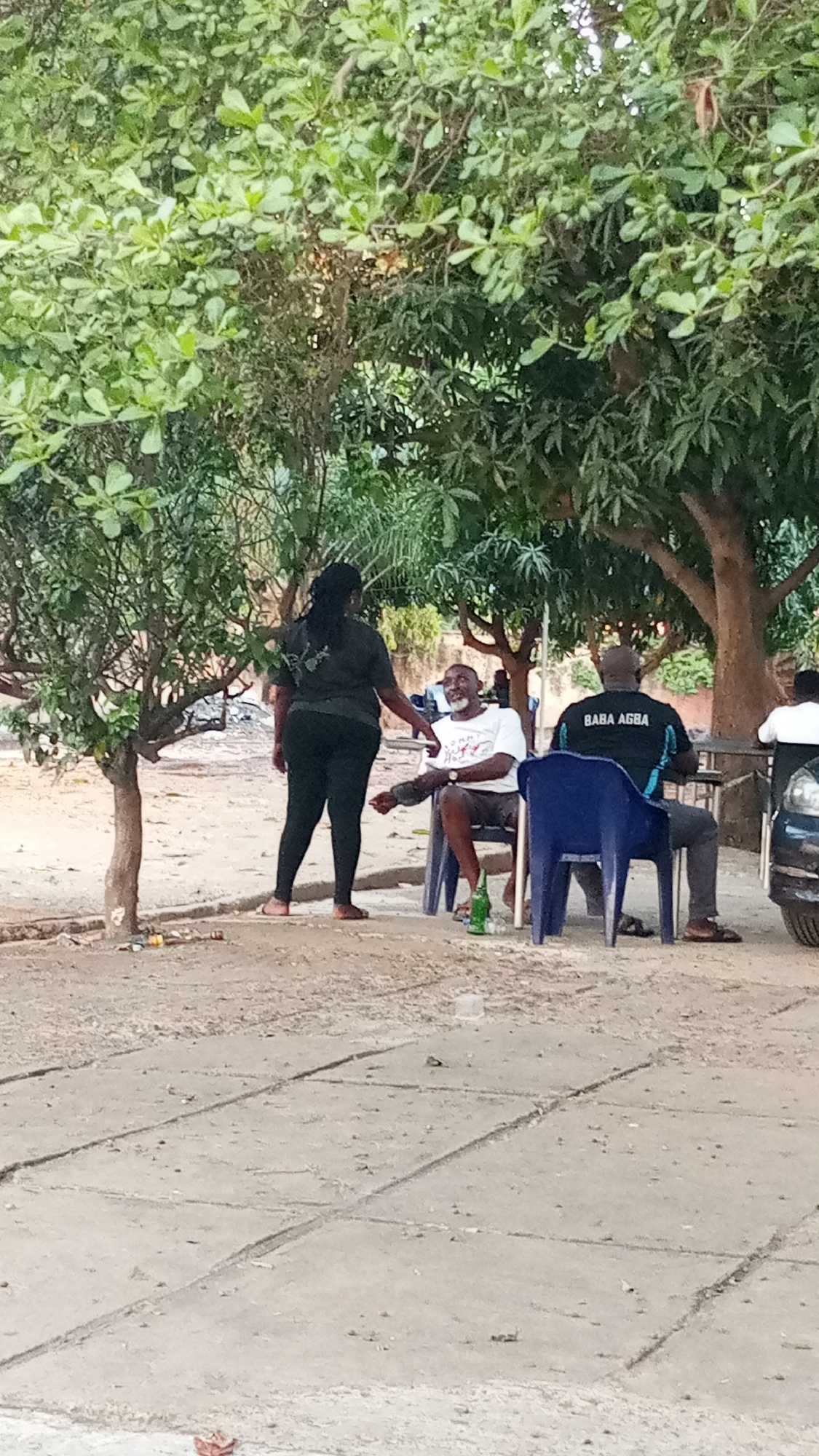 The image depicts an outdoor scene in Adankolo, Nigeria. Three individuals are seated on blue plastic chairs under the shade of trees. The seated figures include one male wearing a white t-shirt and another male with a black t-shirt, labeled "BABA AGBA" in white lettering. A third individual is standing near the first male. A green bottle is on the ground next to the first male. A portion of a parked car is visible on the right side of the image.