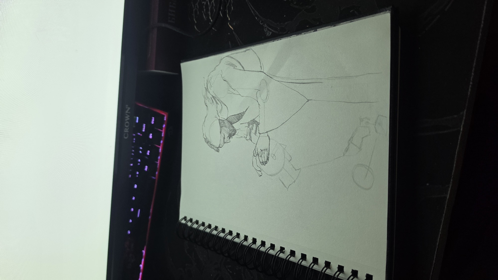 The scene is an indoor workspace, located in the city of Unknown, Unknown. A white, spiral-bound sketchpad is centrally positioned on a dark desk surface. The sketchpad is open, displaying a pencil line drawing on its right page. The drawing portrays two figures. The upper figure, an adult male with a beard and flowing hair or head covering, is depicted in profile, facing left, with his head bowed and body bent forward. He wears a robe or long garment and has his hands positioned over the lower figure. The lower figure is partially sketched, showing a circular shape resembling a head or object, with unfinished lines suggesting a body or base.
Behind the sketchpad, a black mechanical keyboard with glowing pink and purple key backlighting is visible. A computer monitor, featuring the brand 'CROWN' on its bezel, displays a uniform bright white screen. The desk surface is dark. A dark, rectangular object with faint, illegible text is partially visible on the far right background.