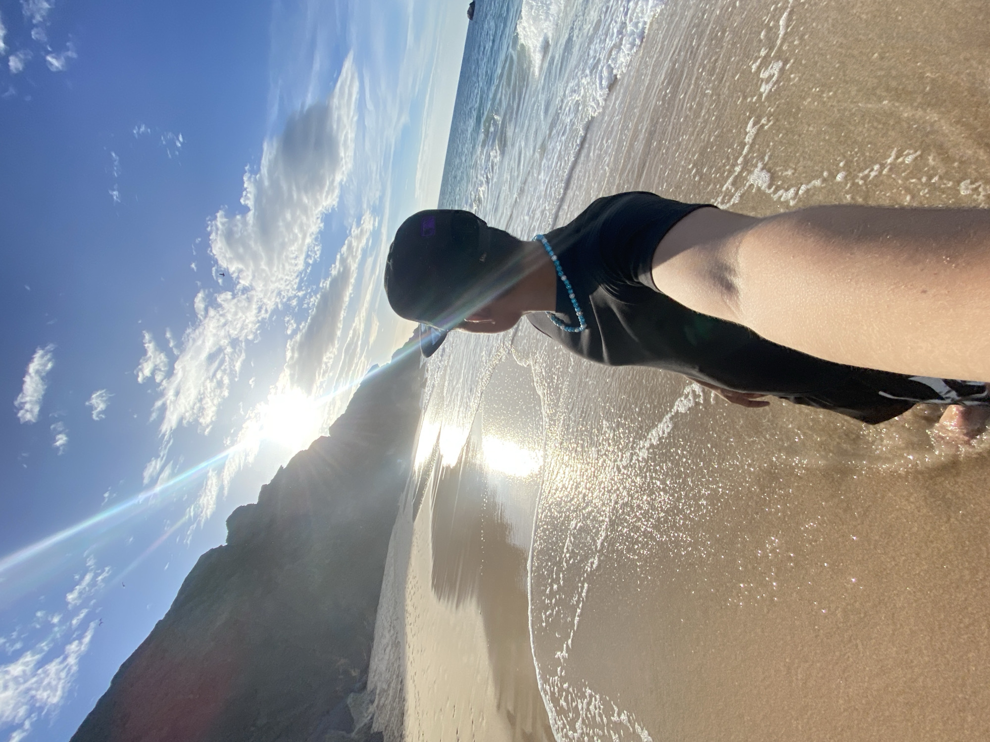 The image depicts a person standing on a beach, looking out at the ocean. The sun is shining brightly, casting a glare across the water and sand. A rocky cliff formation is visible on the left side of the frame. The person is wearing a black t-shirt, black shorts, and a black baseball cap. They are also wearing a turquoise beaded necklace. The water is blue and appears to be calm with gentle waves. The sand is wet near the water's edge and dry further up the beach. The sky is blue with scattered white clouds. The location is identified as the city of Altagracia, Venezuela.