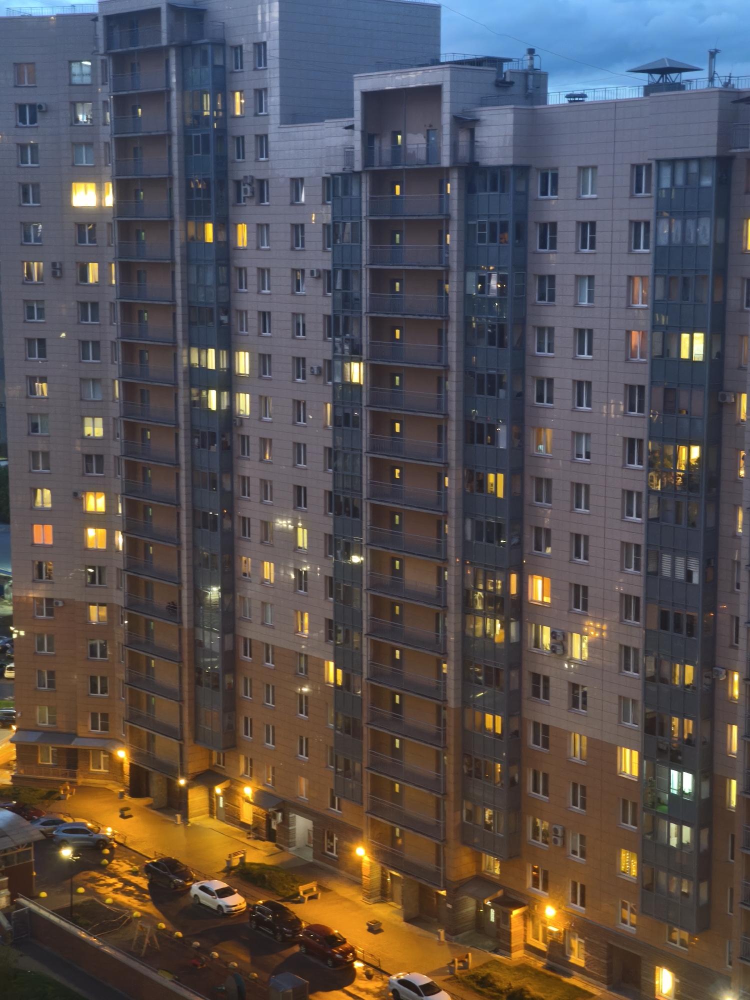 The image shows a tall apartment building complex in Saint Petersburg, Russia at dusk. The buildings have a modern design with light-colored facades and darker grey accents, including glass-enclosed balconies. Many windows are lit, indicating occupancy. Streetlights illuminate the ground level, where several cars are parked. The sky is overcast, suggesting an urban environment. There are no people visible in the scene.
