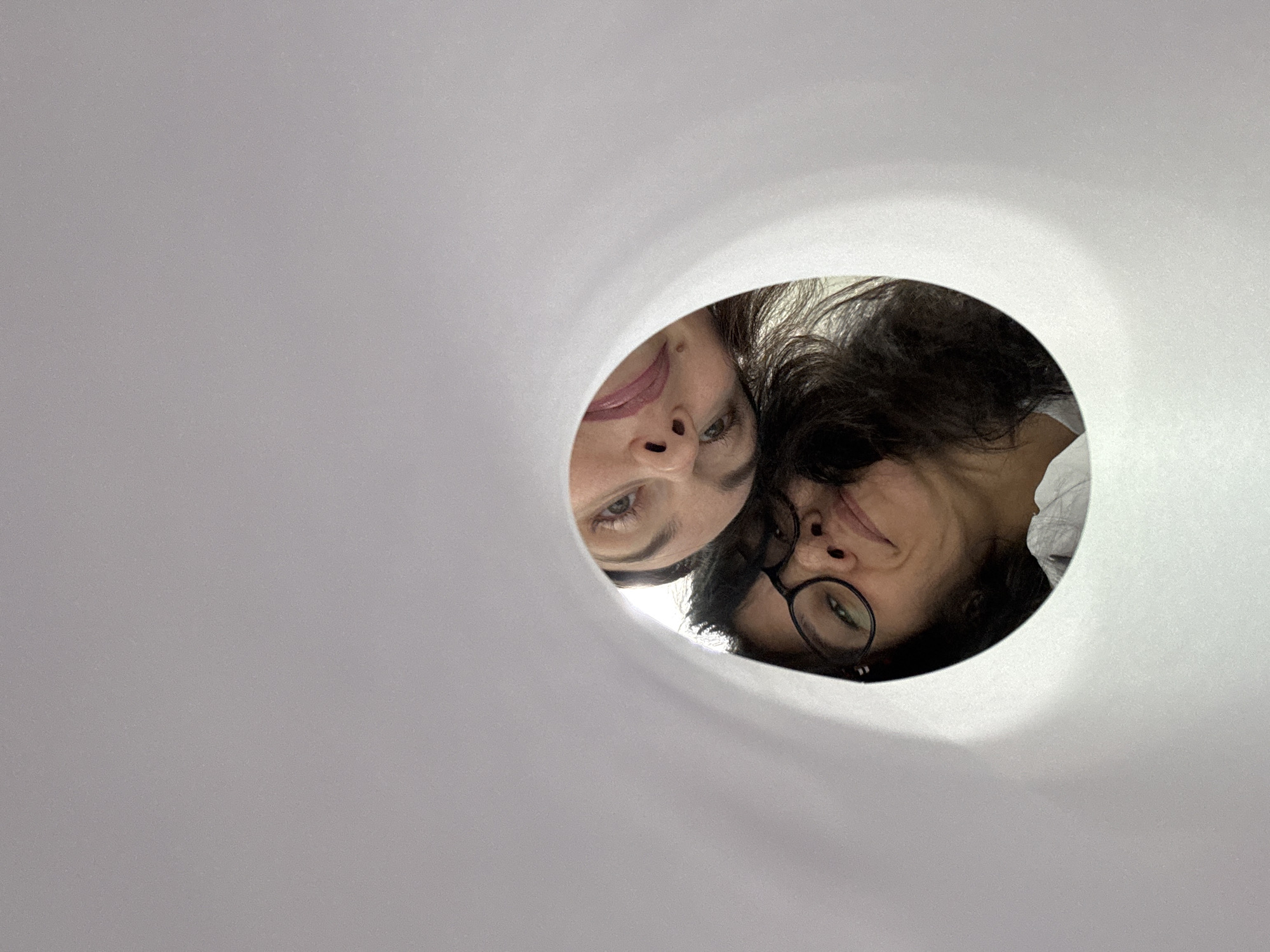 Two people are looking down into the camera through a circular opening in a white surface, possibly a large sheet of paper or similar material. The person on the left has fair skin and dark hair, with visible pink lipstick. The person on the right has darker hair, wears glasses, and is slightly behind the first person. The background is obscured by the white material surrounding the opening. Location is unknown.