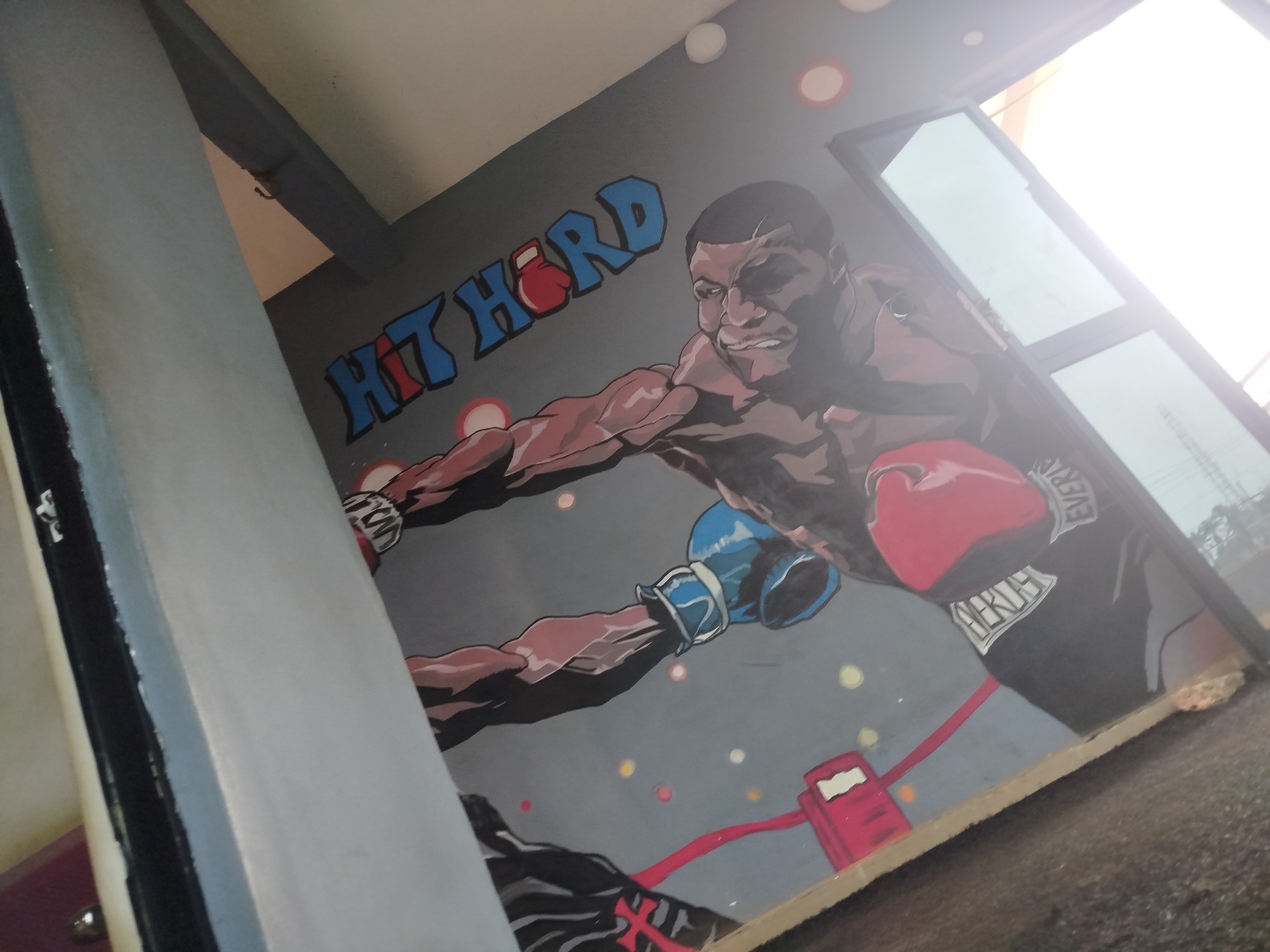 In Uselu, Nigeria, a mural on a grey wall depicts a Black male boxer in mid-punch. The boxer, rendered in a stylized, comic-book art style, wears red boxing gloves on his right hand and a blue boxing glove on his left. He has short dark hair and a determined expression on his face. Above the boxer, the words "HIT HARD" are painted in large, blue, block letters, with a red boxing glove filling the "O". The background of the mural is grey with scattered red and white circular dots, resembling lights or impacts. Below the boxer, a red line connects to a red object that resembles a punch bag or a training dummy. The mural appears to be located in an indoor or semi-outdoor space, possibly a gym or training facility, as indicated by the presence of a large glass window or door on the right side of the image, reflecting bright sunlight. A structural beam runs diagonally across the upper left portion of the image, partially obscuring the top of the mural. The lower edge of the mural is visible, resting on a roughly constructed surface, possibly a concrete base. The overall scene suggests a vibrant and motivational atmosphere within a sports-oriented environment.