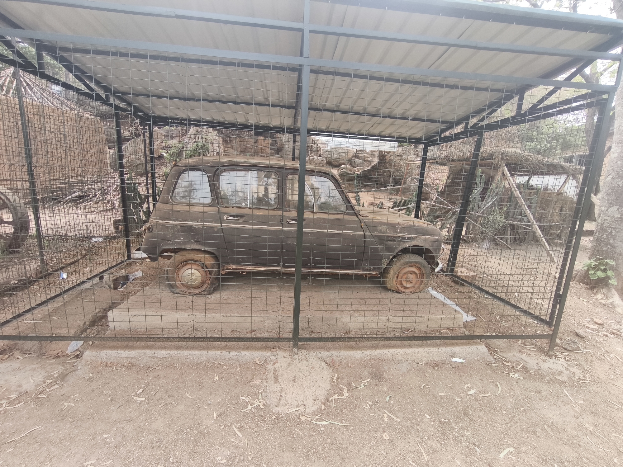 The image shows a dark-colored, rusty Renault 4 car encased in a wire cage with a metal roof structure in what appears to be an outdoor environment. The car is parked on a concrete platform inside the cage. The surrounding environment consists of dirt ground, sparse vegetation, and a backdrop of natural rock formations and other structures, possibly huts or shelters. The location is the Jos Wildlife Park in Jos, Nigeria, as the Renault 4 was the first car ever used by the Park's management.