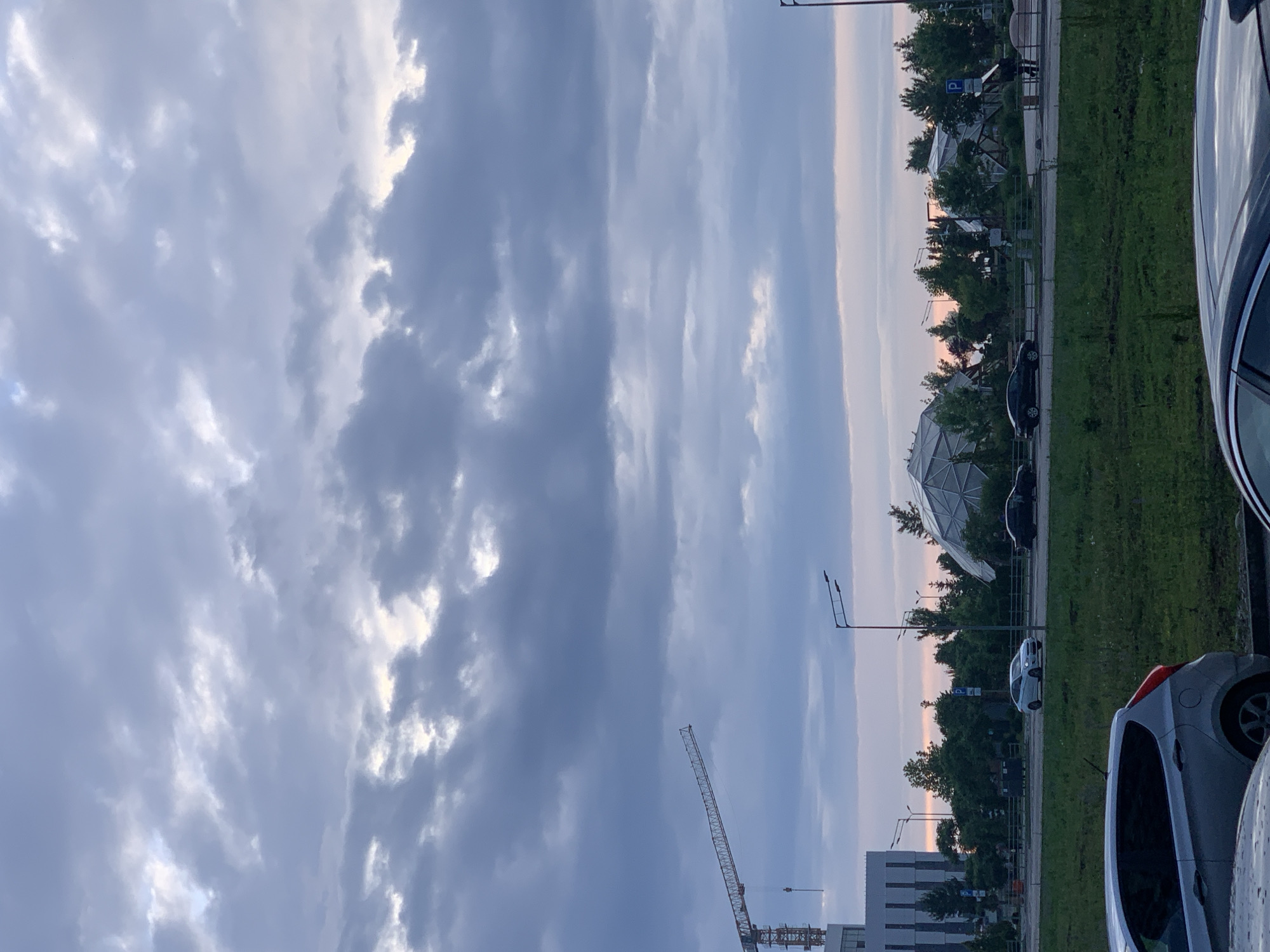 The image depicts a cityscape at dusk with a dramatic, cloudy sky. In the foreground, parked cars are partially visible. A road runs through the mid-ground, with several cars driving on it. Beyond the road, a grassy area is bordered by trees. In the background, there is a modern building with a crane in front of it on the left side of the frame. Several unique, geometric structures resembling domes or pavilions are visible amongst the trees. The overall scene suggests an urban or suburban environment during twilight hours.