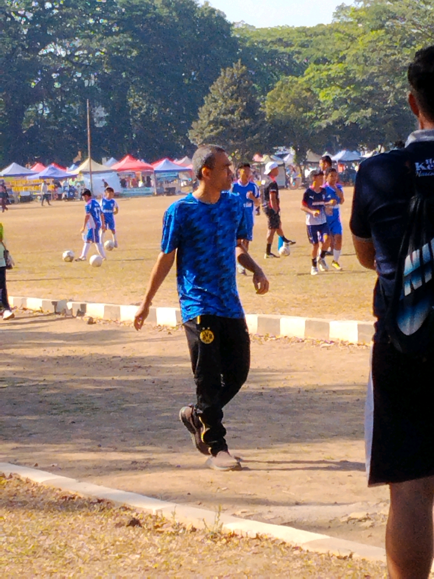 The image depicts an outdoor scene, likely a sports field or park, under bright sunlight. In the foreground, a man wearing a blue patterned t-shirt and black pants is walking away from the camera, facing left. He has short, dark hair and appears to be in motion. Behind him, several other individuals, including children in soccer uniforms, are present on a dirt field. Some of the children are interacting with soccer balls. In the background, there are tents set up, suggesting a possible event or market. Tall trees line the perimeter of the field, and the overall environment appears to be a public space. Based on the context, this scene could be at the Lapangan Bola Rampal, a well-known public sports field in Malang, Indonesia, which often hosts sporting events and community gatherings.