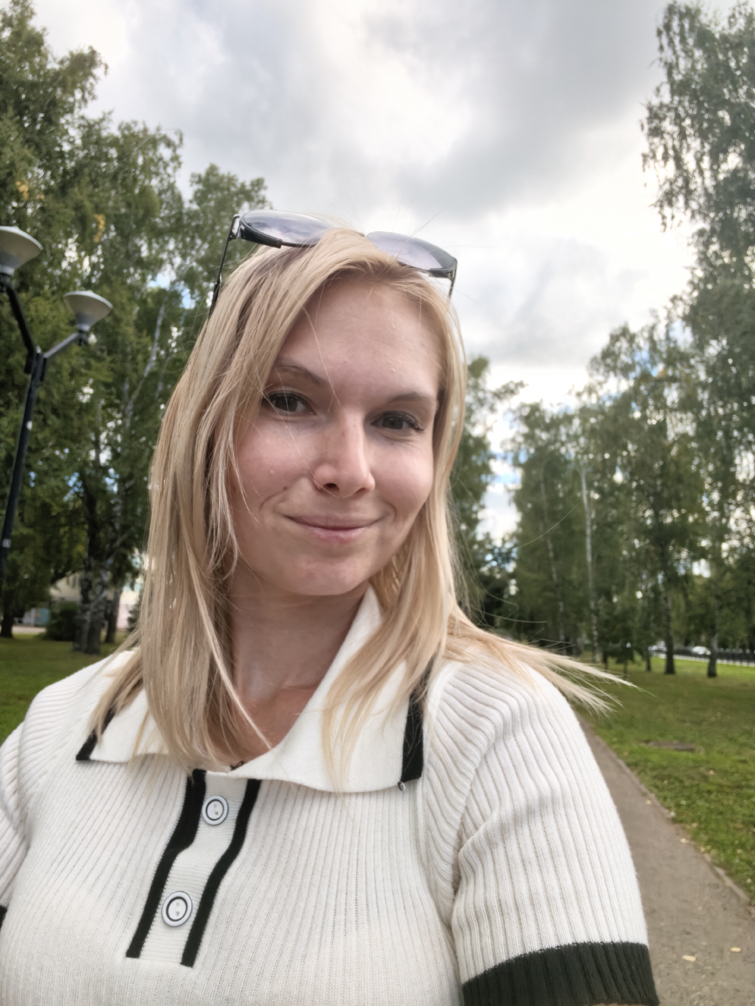 The image shows a woman with blonde hair and sunglasses on top of her head, smiling at the camera. She is wearing a white ribbed sweater with black trim and two black and white buttons. The woman is standing outdoors in what appears to be a park or a tree-lined street. In the background, there are trees, a paved path, and a cloudy sky. There are also two street lamps visible on the left side of the image. The setting is consistent with a public area in Sterlitamak, Russia, likely a park or a street with a pedestrian walkway.