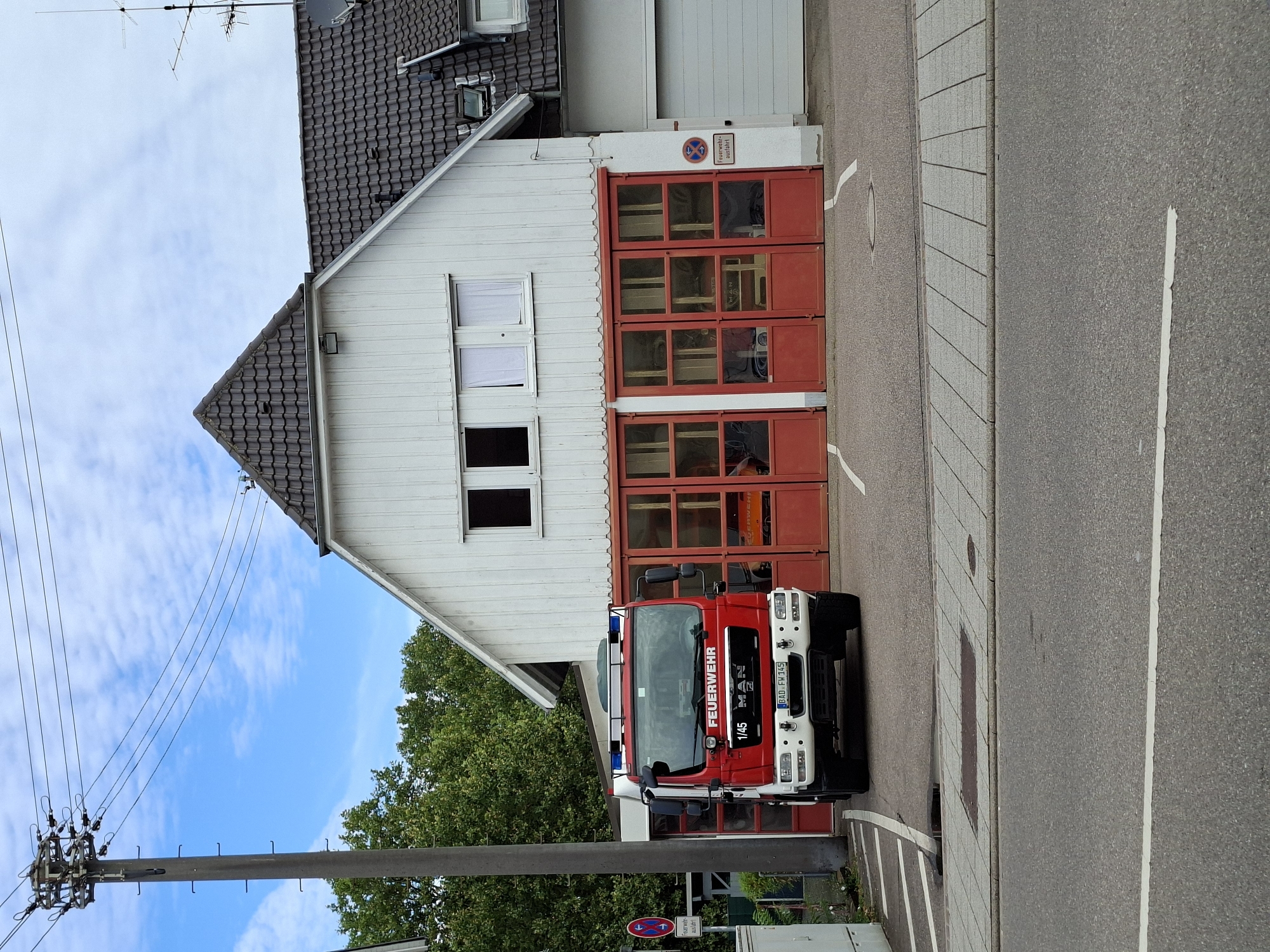 The image captures a fire station in Sinzheim, Germany. A red fire truck, identified by "FEUERWEHR" on its front, is parked in front of the station's entrance. The station building is white with a dark tiled roof and features several large red doors with windows. To the left of the station, a tall utility pole supports multiple overhead wires. In the background, lush green trees frame the scene, and a bright blue sky with scattered clouds is visible. The foreground shows a paved area and a road. A traffic sign with two red arrows is visible near the utility pole.