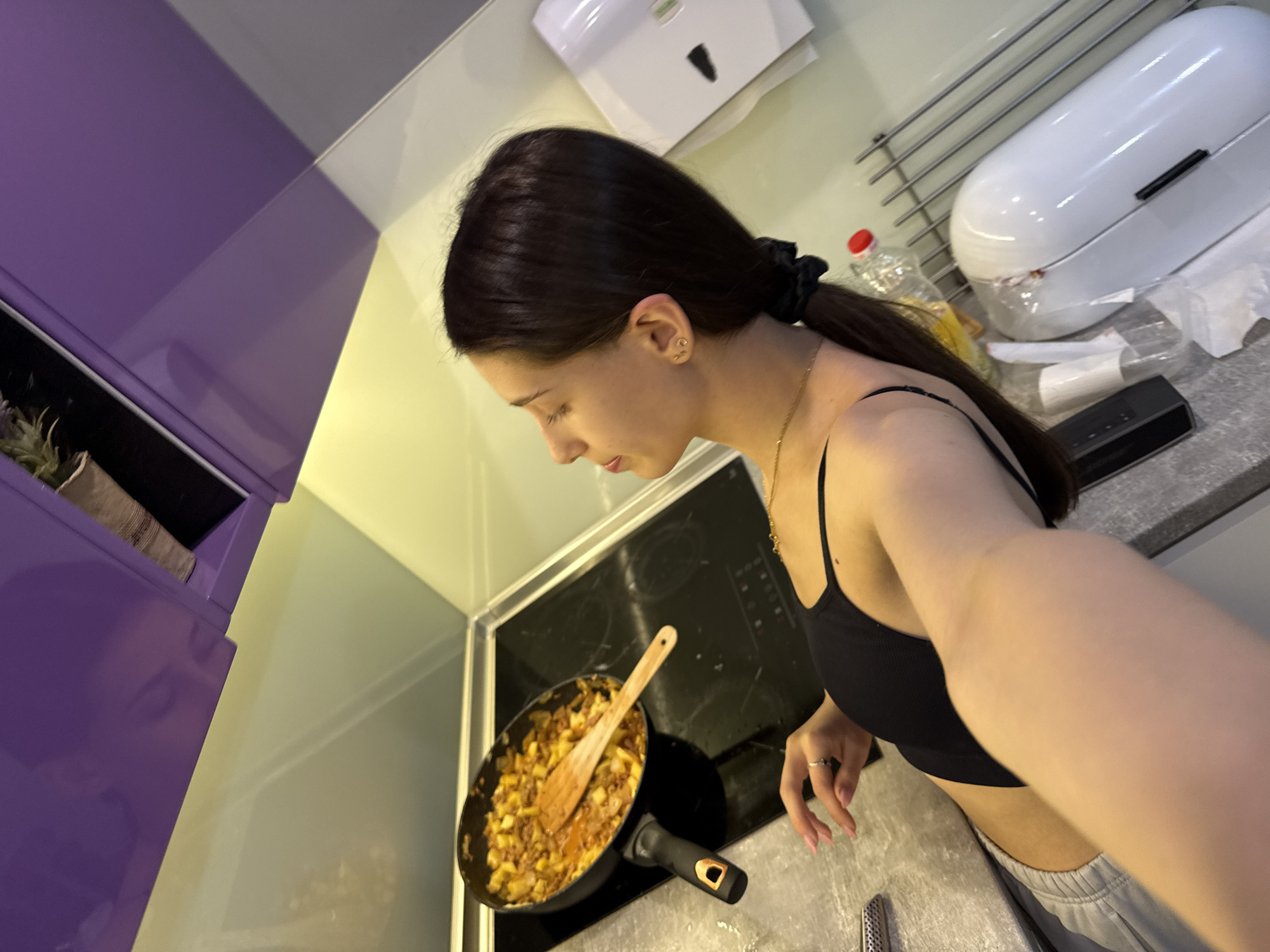 A young woman with dark hair pulled back in a ponytail is stirring food in a pan on a stovetop. The pan contains cooked potatoes and what appears to be meat. She is wearing a black tank top and grey sweatpants. The kitchen features a purple cabinet, a white paper towel dispenser, and a white breadbox. The location is likely a domestic kitchen in Sofia, Bulgaria, but there is no definitive evidence to confirm this.