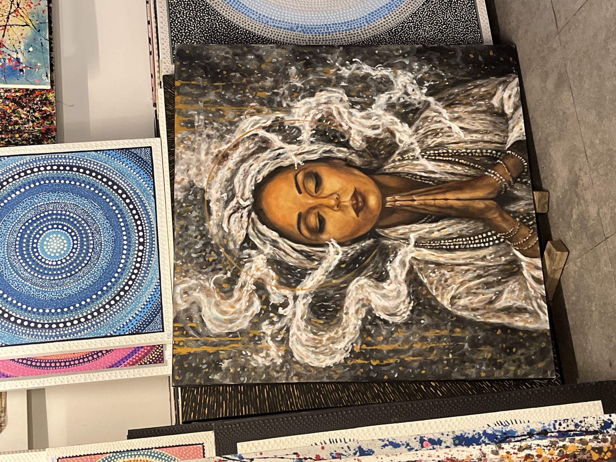 The image depicts a collection of paintings, likely at a market or art exhibit. The main focus is a painting of a woman with closed eyes, fair skin, and flowing white hair. She appears to be lying down, with her hands clasped in a prayer-like position. She is adorned with beaded jewelry. The background of this painting is dark with gold accents. To the left, there is a painting with a concentric circle pattern in shades of blue and white. Other paintings are visible in the background and foreground, including ones with abstract patterns. Based on the information provided, the location is Kuta Selatan, Indonesia.