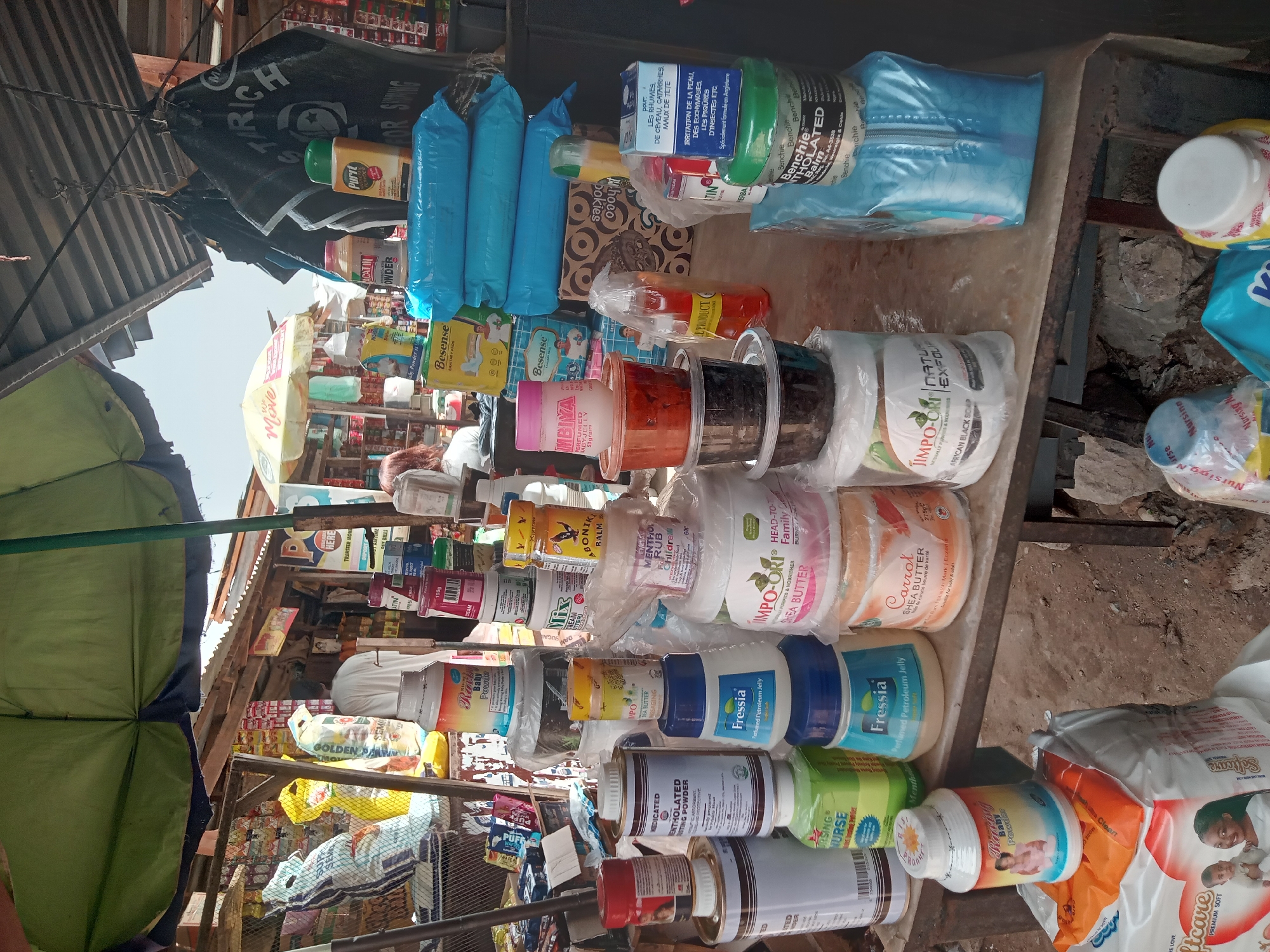 The image shows a stall at a market in Sabon Tasha, Nigeria. The stall is under a makeshift awning of green and dark fabric. Various personal care and cosmetic products are displayed on a surface, including lotions, creams, powders, sanitary pads, and balms. Brands such as "Besense," "Impo-Ori," "Fressia," "Purit," and "Benchie" are visible. Some products are in containers of varying sizes and shapes, while others are in blue plastic packaging. In the background, there are more goods, shelves, and possibly other market stalls. The lighting is bright, suggesting it is daytime.