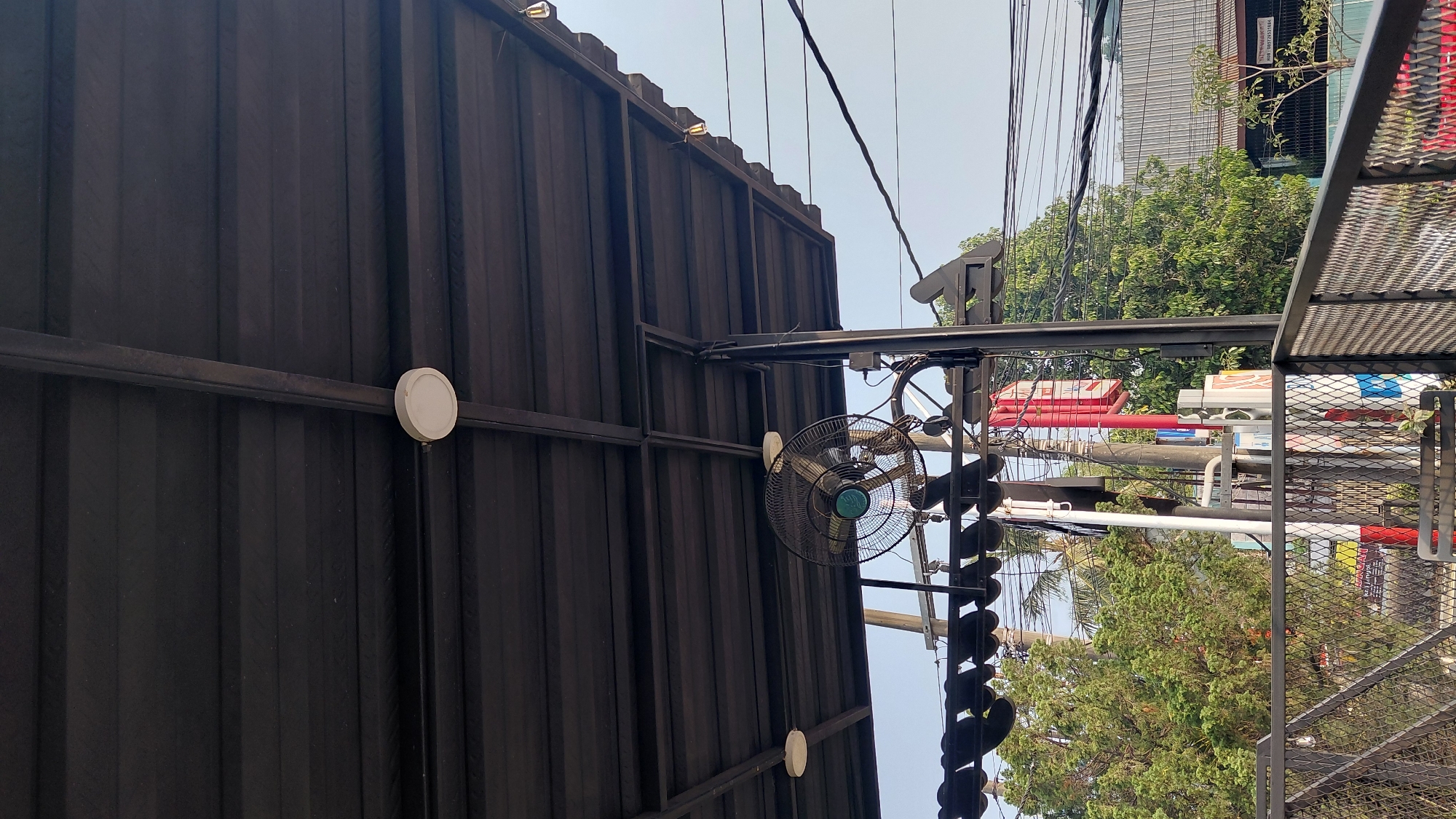 The image captures an outdoor area beneath a dark corrugated metal roof supported by black metal beams and posts. A black oscillating fan with a green central component is mounted beneath the roof, angled downwards. Two circular white lights are also attached to the underside of the roof. On the right side of the image, a portion of a black metal fence with a diamond pattern is visible, extending into the foreground. Beyond the fence, lush green trees are prominent, with power lines and utility poles interspersed among them. In the background, a building with a silver roof and a red sign with white text is partially visible. The sky is a clear, light blue. The scene appears to be an open-air seating area or walkway, possibly at a restaurant or cafe, with elements suggesting an urban or suburban environment. There are no people visible in the image. This specific view is likely from a public space in or around Karanglewas, Indonesia, given the context provided.