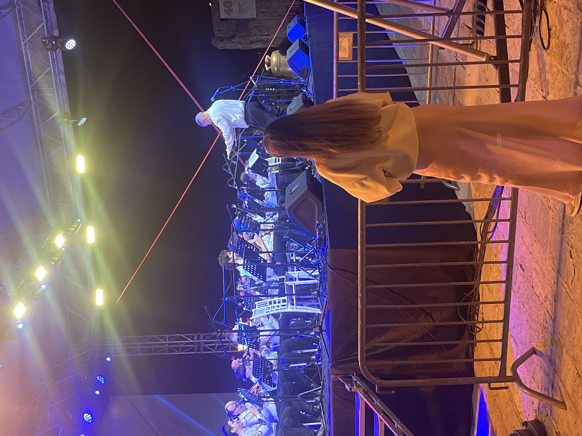 At nighttime in Budva, Montenegro, an outdoor concert unfolds on a brightly lit stage. An orchestra is visible on the stage, with musicians seated and facing a conductor who leans toward them. The stage is illuminated with a combination of blue and yellow lights, with a red laser beam visible in the upper-left corner. A person wearing a light-colored jacket and long hair, appears to be a spectator, standing near a metal fence on the right side of the frame, observing the performance. The background beyond the stage is dark, suggesting the concert is occurring in the evening.