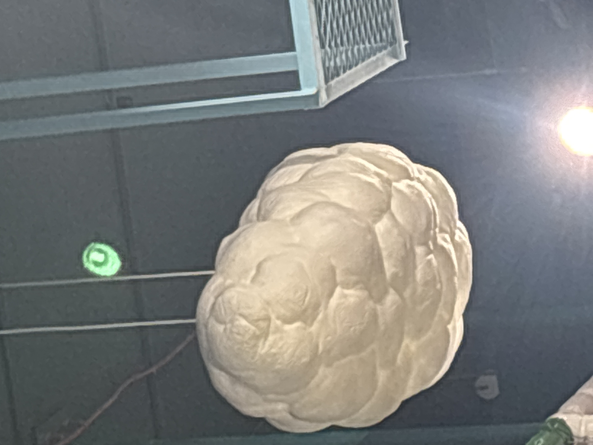 The image depicts a large, white, cloud-shaped object suspended from the ceiling. The object has a textured surface with numerous folds and indentations, resembling a stylized cumulus cloud. It is illuminated by overhead lighting, casting soft shadows on its surface. To the left of the cloud object, a thin, dark wire is visible, also suspended from above. Above and slightly to the right of the cloud, a small, circular, green light source is illuminated. In the background, a section of a light blue, metal grid structure is visible, possibly part of the building's architecture or a fixture. The overall environment appears to be indoors with a dark ceiling. The context of Dubai, United Arab Emirates, is not visually evident in this image.