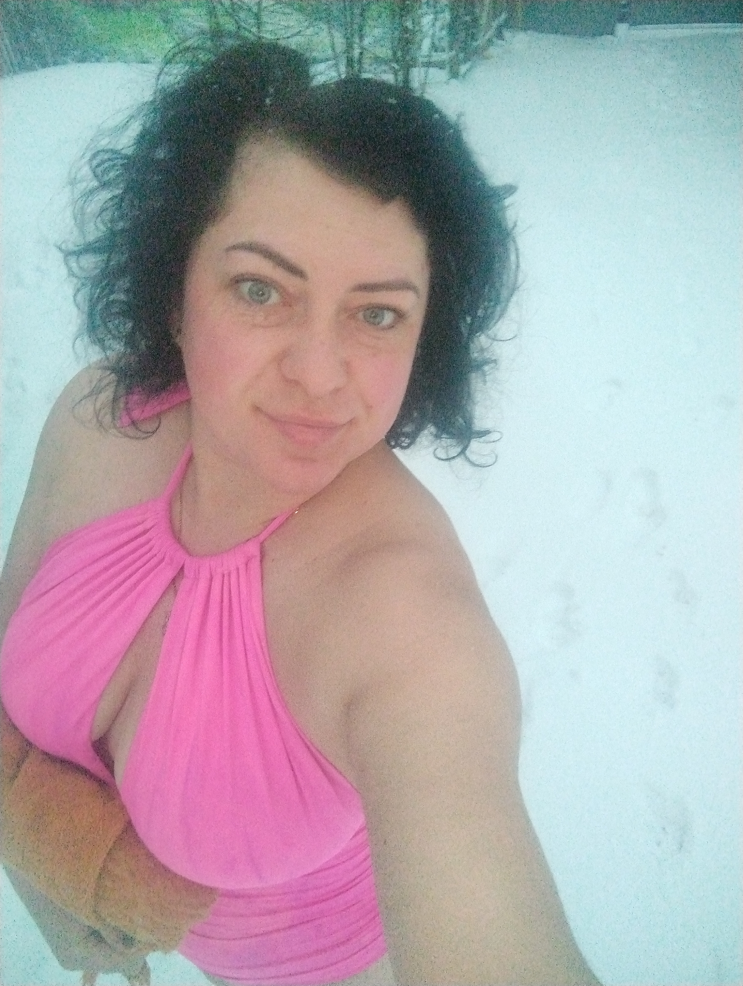 A woman with dark curly hair and light eyes stands in a snow-covered area, wearing a pink swimsuit. Her right arm is raised, possibly taking a selfie. She appears to be holding something tan-colored with her left hand, pressed against her torso. The background shows indistinct vegetation and a snow-covered landscape. While the prompt mentions Grishanki, Russia, this location cannot be verified from the image alone.