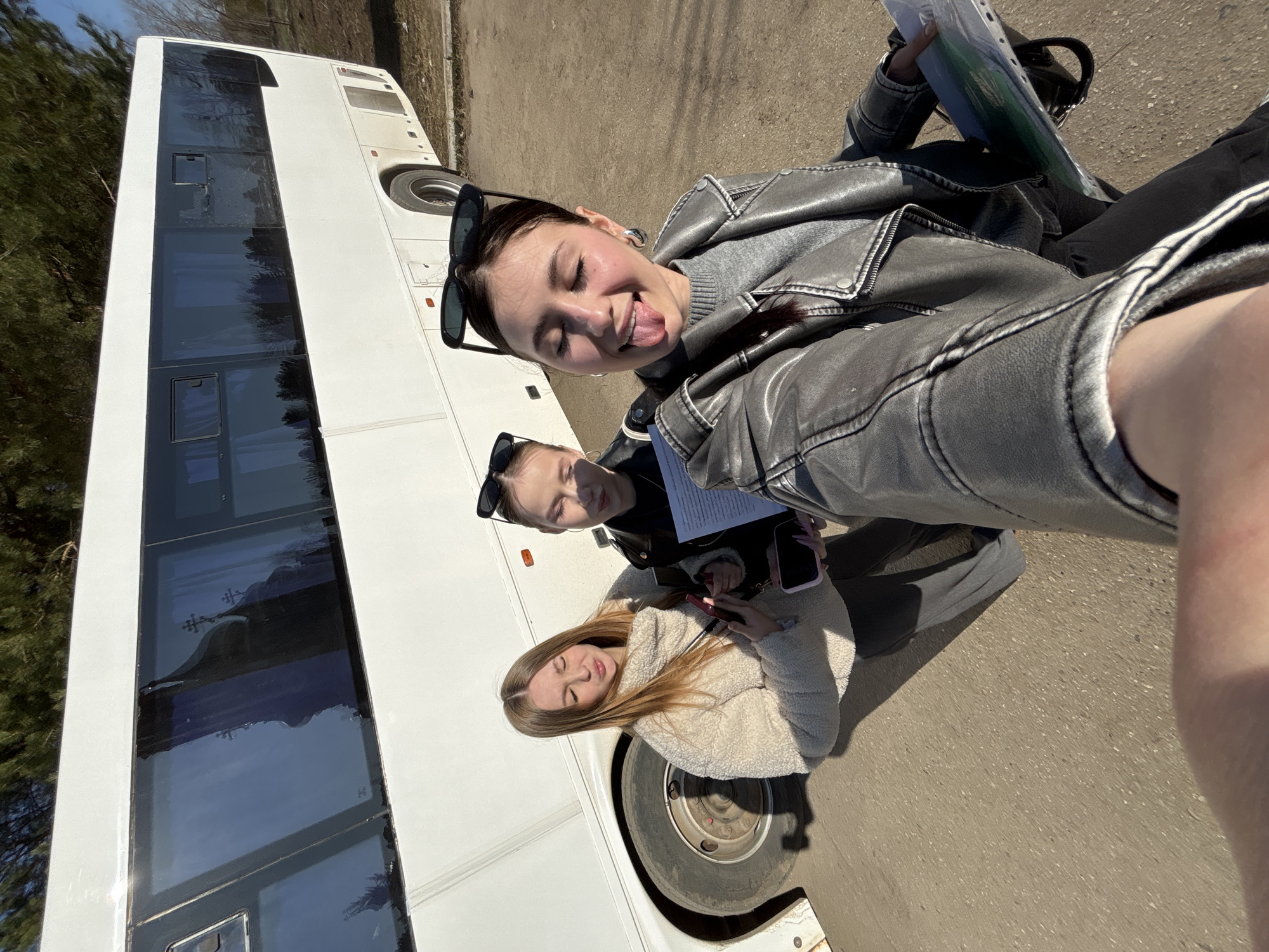 A sideways outdoor photograph captures three young women posing next to a large white coach bus on a bright, sunny day. The woman closest to the camera, wearing a gray leather jacket and sweater, is taking a selfie, playfully sticking her tongue out with her eyes slightly closed. Black sunglasses rest on top of her head. Behind her, another woman with similar sunglasses covering her eyes, also wearing a dark jacket, looks towards the camera while holding some papers. Further back and lower in the frame, a third woman with long light brown hair, wearing a cream fuzzy jacket, looks towards the camera with her eyes partially closed, holding a pink cell phone.
The large white bus dominates the left side of the frame, with its dark, tinted windows reflecting the bright sky. The women are positioned on an asphalt or concrete surface next to the parked bus. Strong sunlight casts clear shadows, indicating midday or early afternoon. In the far left background, some trees with bare branches are visible. The casual jackets worn by the women suggest mild weather. The scene portrays a casual outdoor gathering, likely a break during a bus journey in Chaikovsky, Russia. No clearly legible text is visible.