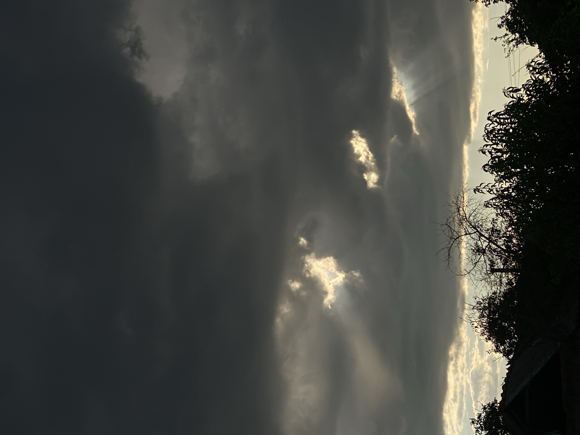 The image depicts a dramatic sky with heavy, dark clouds, characteristic of an approaching storm or the aftermath of one. Rays of sunlight pierce through gaps in the clouds, creating a striking contrast and illuminating portions of the sky with a golden hue. Silhouetted against the sky are the dark shapes of trees and what appears to be the roofline of a building, likely in a rural or suburban setting. The scene is set in Cairaclia, Moldova. There are no people or specific actions depicted in the image. The environment is dominated by the atmospheric conditions of the sky and the dark silhouettes of the foreground elements.