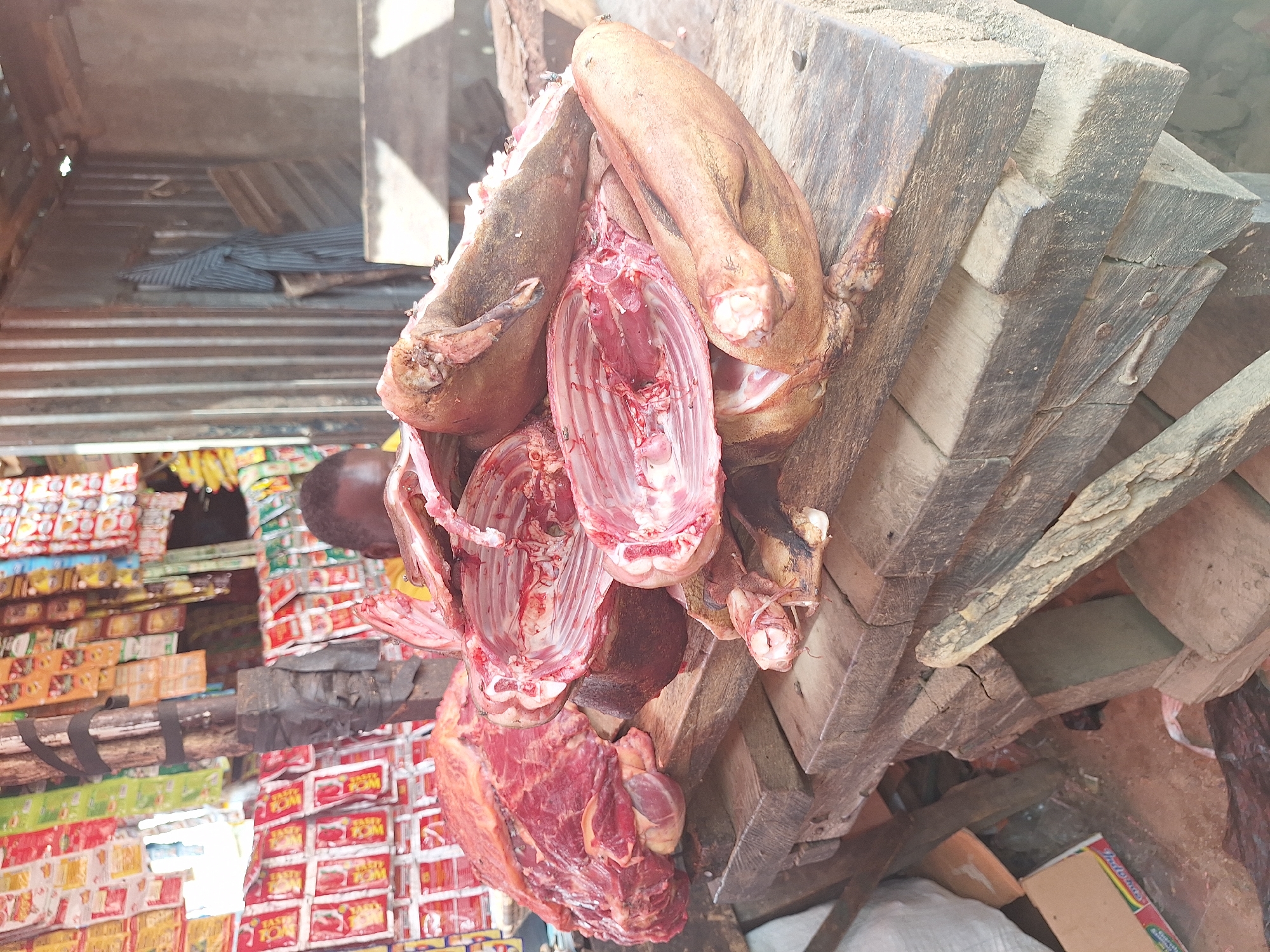 The image depicts a butcher's stall in Sabon Tasha, Nigeria. Various cuts of meat, including what appears to be pork, are hanging from a wooden structure. The meat is exposed to the open air. Behind the stall, there are stacks of packaged goods, likely other food items for sale. The overall scene suggests a typical open-air market environment. A person can be seen in the background behind the meat.