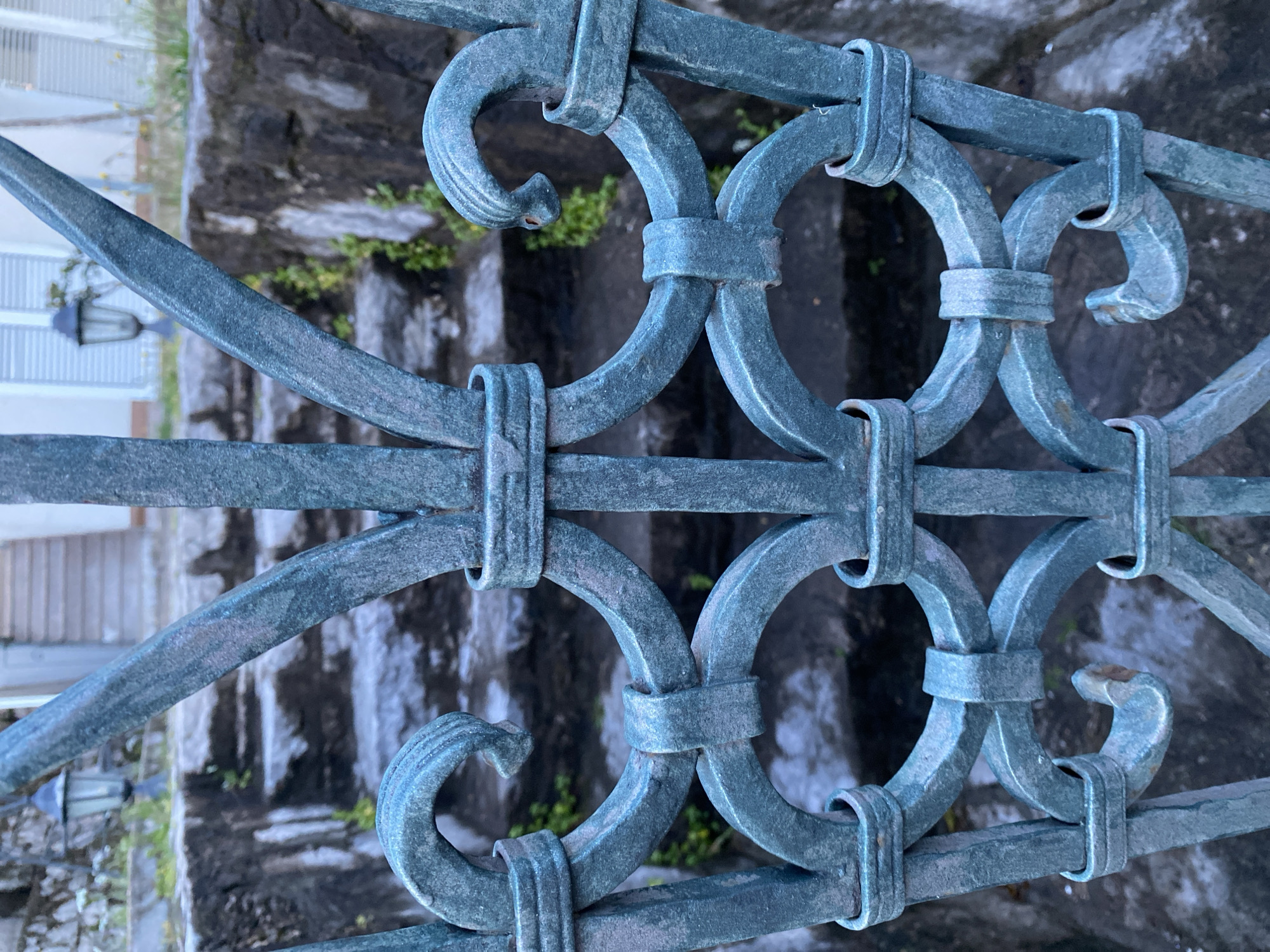 An eye-level, medium-close-up shot shows a section of an ornate, aged metal railing against a backdrop of stone steps and vague architectural elements, potentially in Budva, Montenegro. The railing, painted in a muted teal or blue-green hue, exhibits a repeating pattern of interconnected circles and decorative curls. Horizontal bars run across the design. The metal surface shows signs of age, with slight variations in color and texture indicating weathering or oxidation.
Behind the railing, a stone staircase ascends, visible in a blurry state. Patches of moss or small plants grow in the crevices of the stone steps. In the background, partly visible through the railing, are the blurred forms of buildings and a black lamppost. The light suggests it is daytime with potentially diffused sunlight.