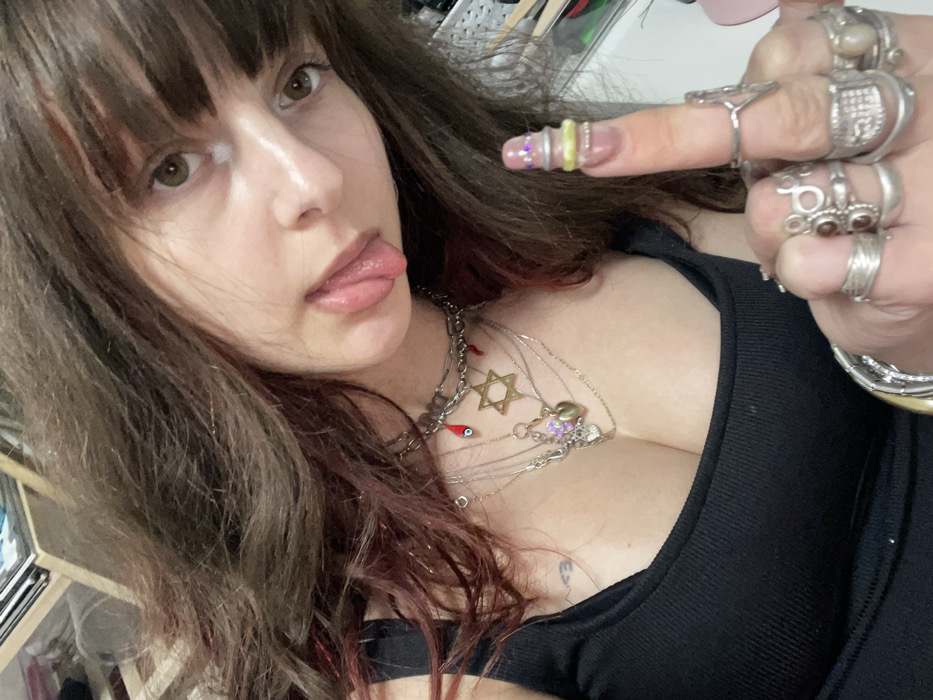A young woman with brown hair and bangs is sticking out her tongue. She is wearing a black tank top and multiple silver necklaces, including one with a Star of David pendant. She is wearing many silver rings on her right hand. Her nails are decorated with yellow bands and rhinestones. The overall composition is a selfie taken from a low angle.