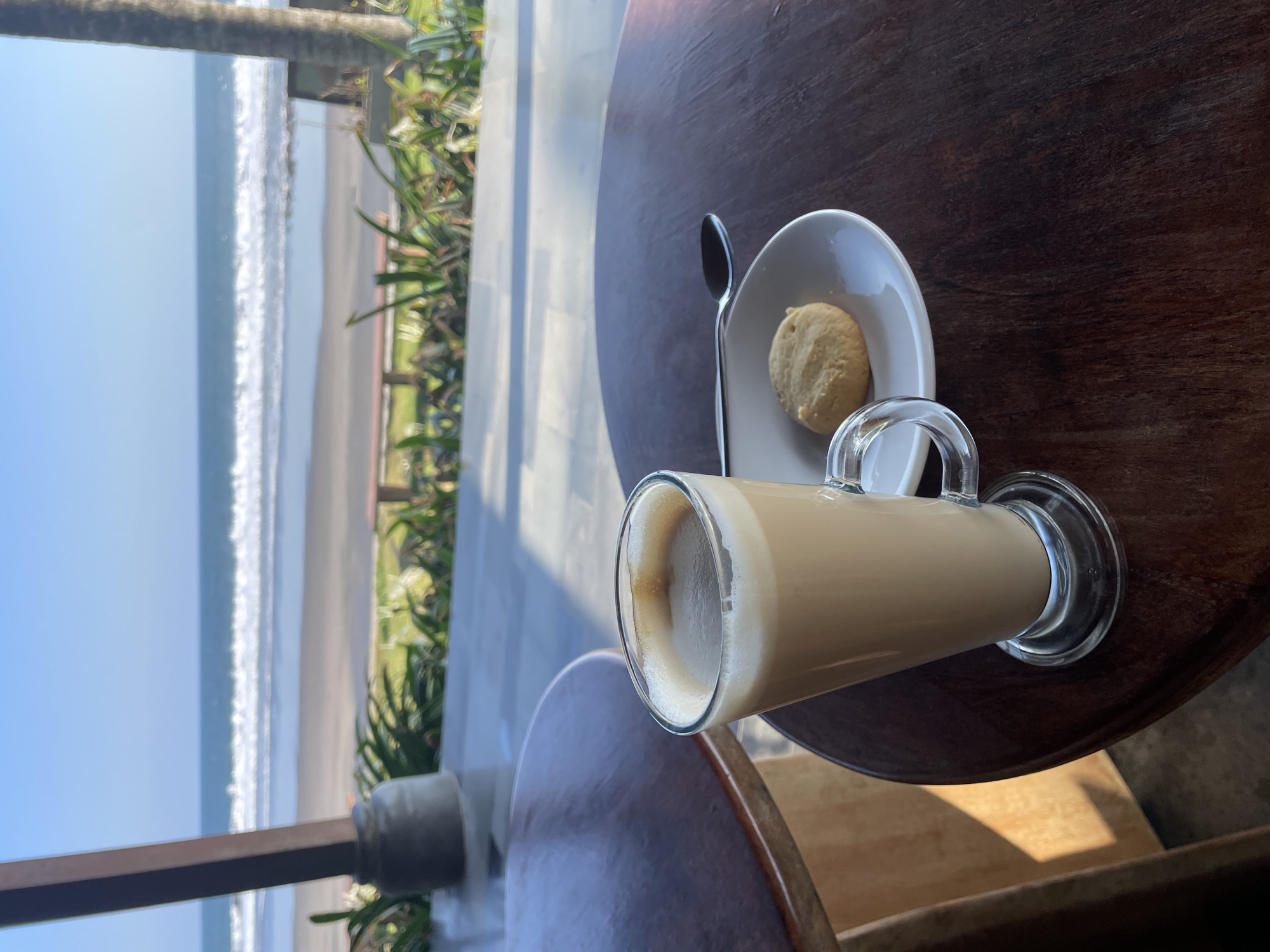 A latte in a tall glass with a cookie on a saucer with a spoon sits on a wooden table. The table is set on a patio with a view of the ocean in Selemadeg Timur, Indonesia. The ocean has waves crashing on the shore. There are palm trees and green foliage visible. The sky is clear and blue.
