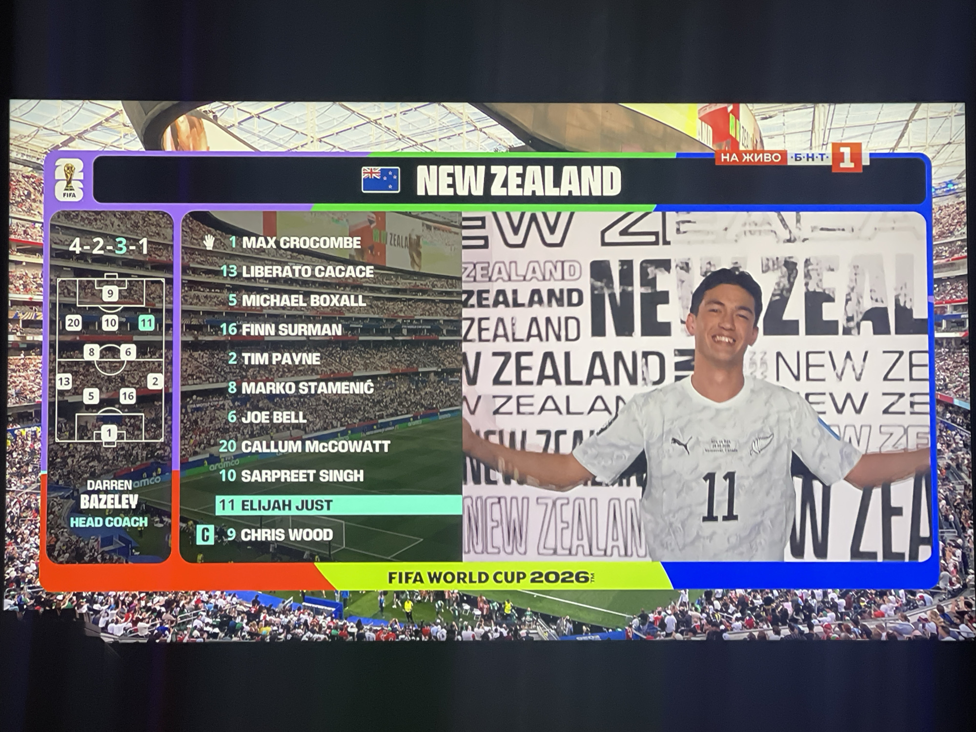 The image displays a screen showing a soccer match lineup for New Zealand. The screen is divided into sections. On the left, a formation diagram labeled "4-2-3-1" is shown, with player numbers positioned on a soccer field graphic. Below this is the team's head coach, Darren Bazeley. The central section lists the players with their numbers and names, including Max Crocombe, Liberato Cacace, Michael Boxall, Finn Surman, Tim Payne, Marko Stamenic, Joe Bell, Callum McCowatt, Sarpreet Singh, Elijah Just, and Chris Wood. The top banner features the FIFA World Cup logo and the text "NEW ZEALAND" with the New Zealand flag. The right side of the screen shows a player wearing a New Zealand jersey with the number 11, smiling, with the text "NEW ZEALAND" repeated in the background. A green banner at the bottom reads "FIFA WORLD CUP 2026". The image is a photograph of a television screen, with a portion of the stadium and crowd visible beyond the screen. There is no indication of the city of Ruse, Bulgaria in the image.