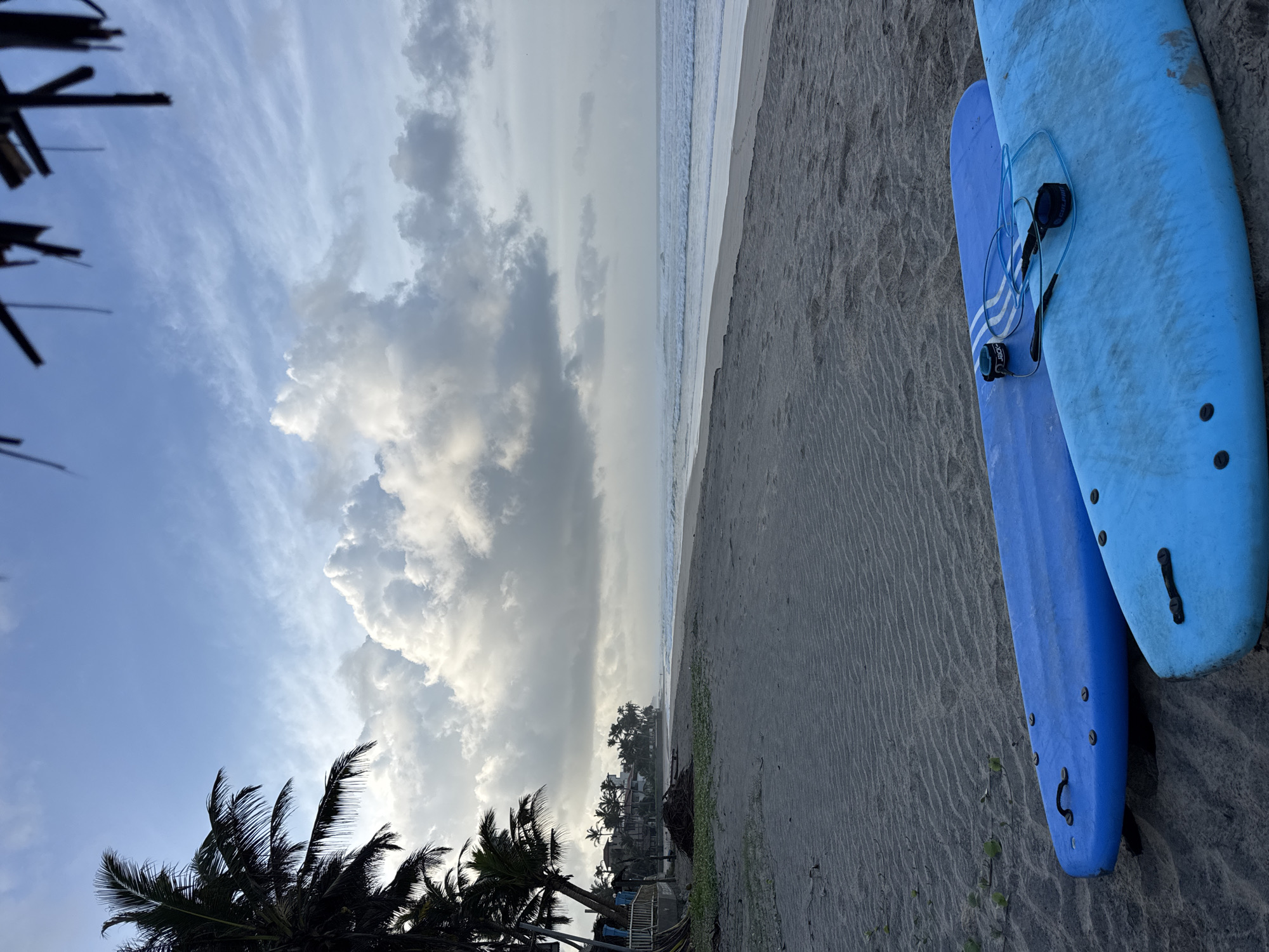 The image depicts a serene beach scene in Kathaluwa, Sri Lanka. Two blue surfboards are laid out on the sandy shore in the foreground. Beyond them, the ocean stretches to the horizon, with gentle waves lapping the coastline. Palm trees line the beach in the background, interspersed with buildings, suggesting a coastal settlement. The sky is partly cloudy, with dramatic cumulus clouds casting shadows on the sea and sand. The overall atmosphere is tranquil, with the muted light indicating either early morning or late afternoon. There are no people visible in the frame.