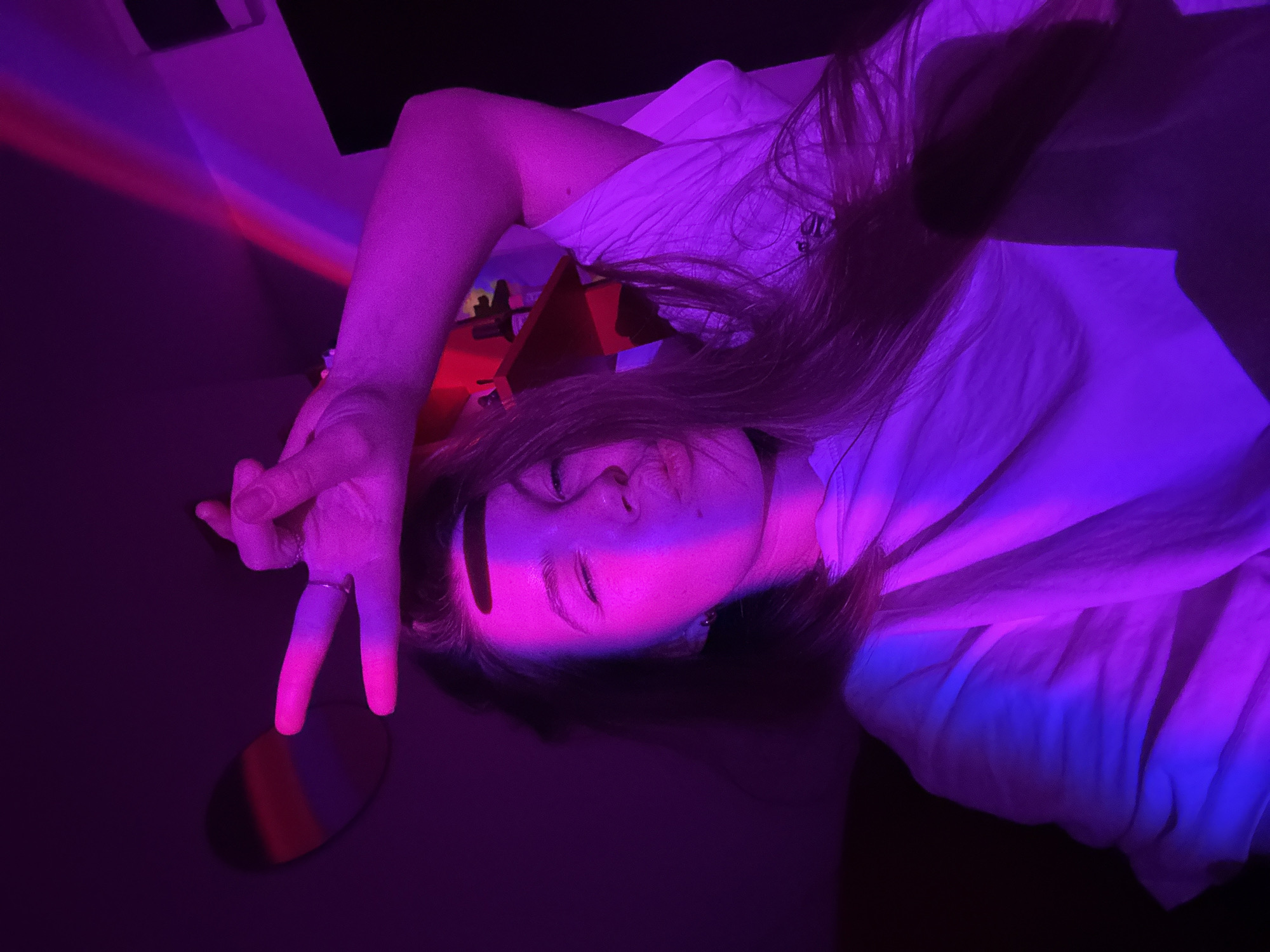 The image displays a person, likely female, captured in a darkly lit indoor setting, bathed in striking neon purple, pink, and blue light. The subject is positioned diagonally, appearing to be lying down, with her head towards the bottom right of the frame. Her face is illuminated by vibrant pink light, highlighting her features, and her eyes are open, looking towards the camera. Long, dark hair cascades around her head.
Her right hand is raised, making a "peace" sign, with two fingers extended upwards. A silver ring is visible on her ring finger. Beneath her hand, an oval-shaped, reflective object catches the colored light, showing streaks of red, blue, and purple. White fabric, possibly a shirt or bed linens, is draped around her upper body and arm, also glowing under the purple illumination. In the upper left background, distinct diagonal streaks of red and blue light are projected onto a surface, hinting at the source of the atmospheric lighting.
The scene suggests an evening or nighttime setting, with artificial colored lights creating a specific, artistic mood, possibly during a creative photoshoot or a relaxed gathering. The overall emotion conveyed is relaxed and perhaps playful due to the hand gesture. There is no visible text. The provided context indicates the location is Vozdovac Urban Municipality, Serbia.